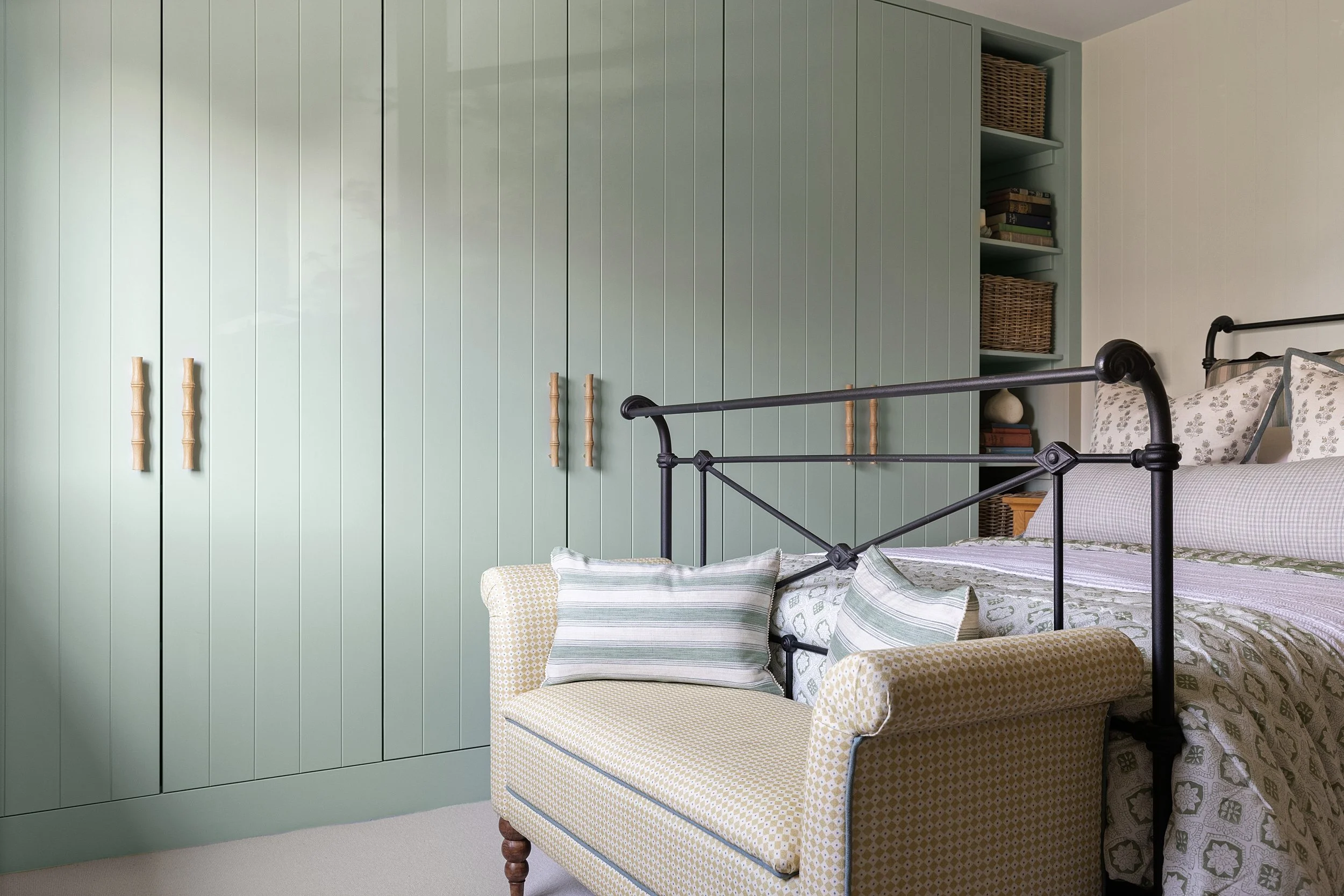A bedroom featuring a green wardrobe with wooden handles, a black metal bed frame, a yellow upholstered bench with pillows, and a built-in bookshelf with wicker baskets and books.