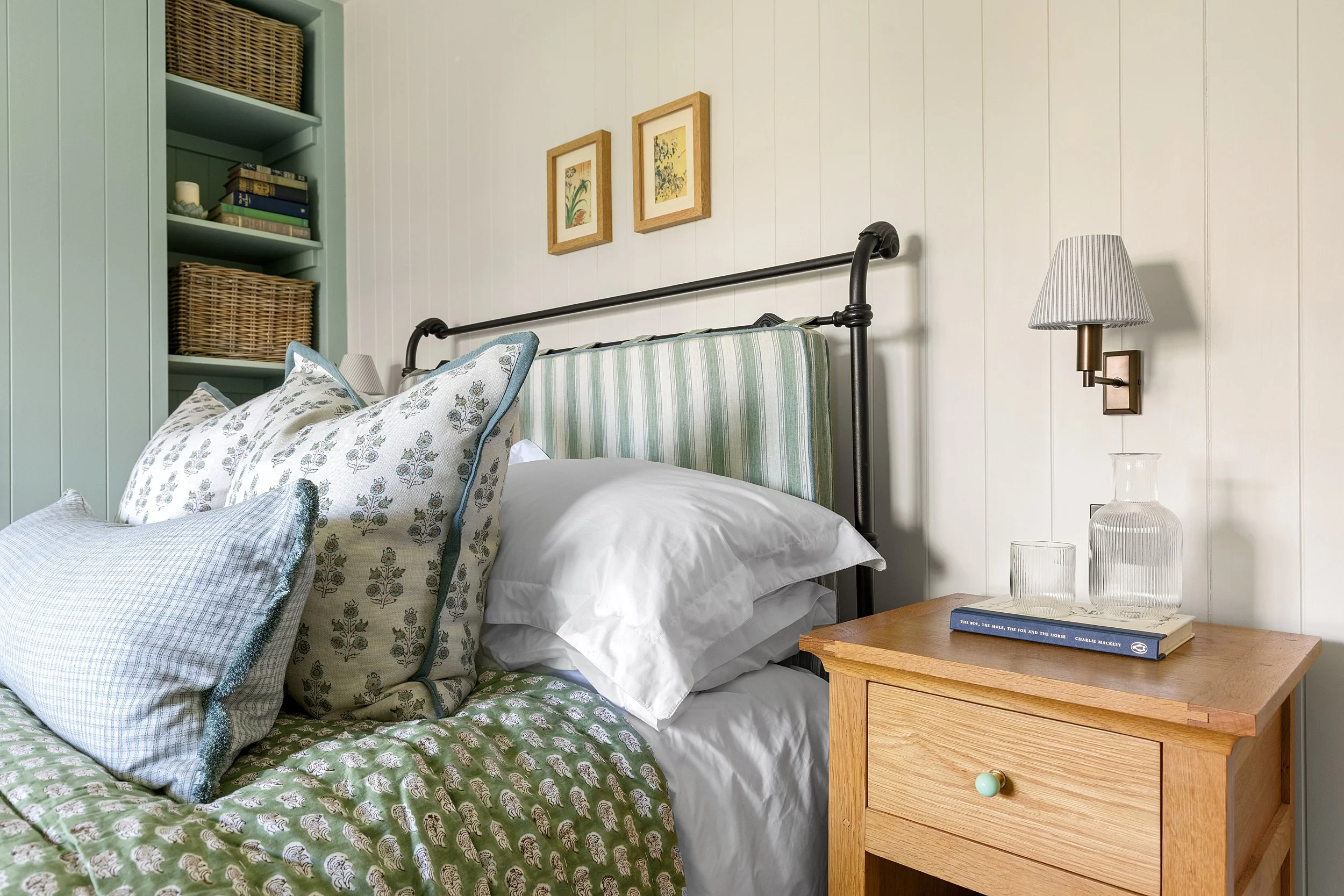A cozy bedroom with a bed featuring a green, patterned quilt and multiple decorative pillows, a wooden nightstand with a glass vase, glasses, and a book, and a wall-mounted lamp next to framed botanical art on a white paneled wall.