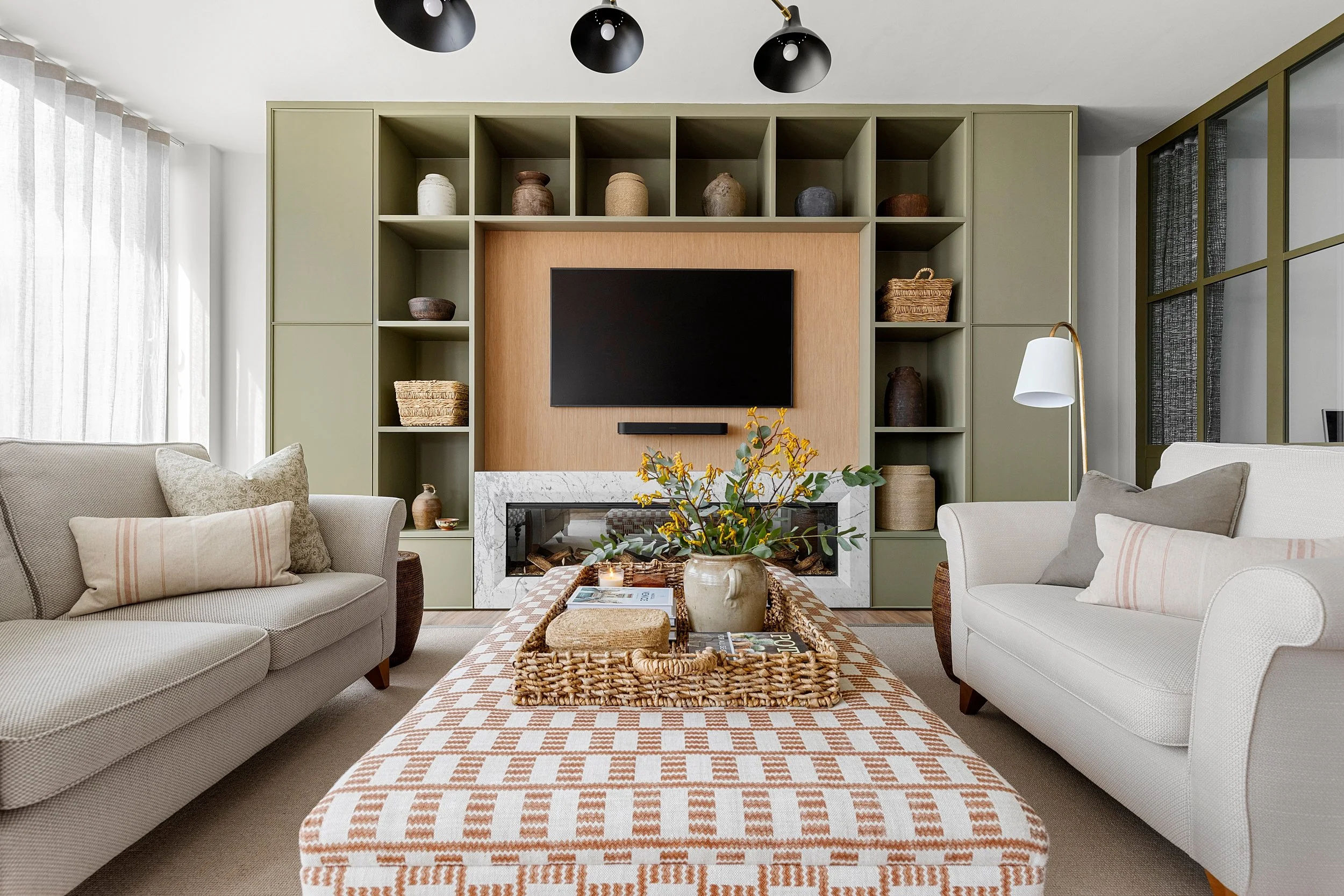 Living room with beige and white sofas, a patterned ottoman with a tray, a vase with flowers, a wall-mounted TV, a fireplace, built-in shelves with decor, and a large window with sheer curtains.