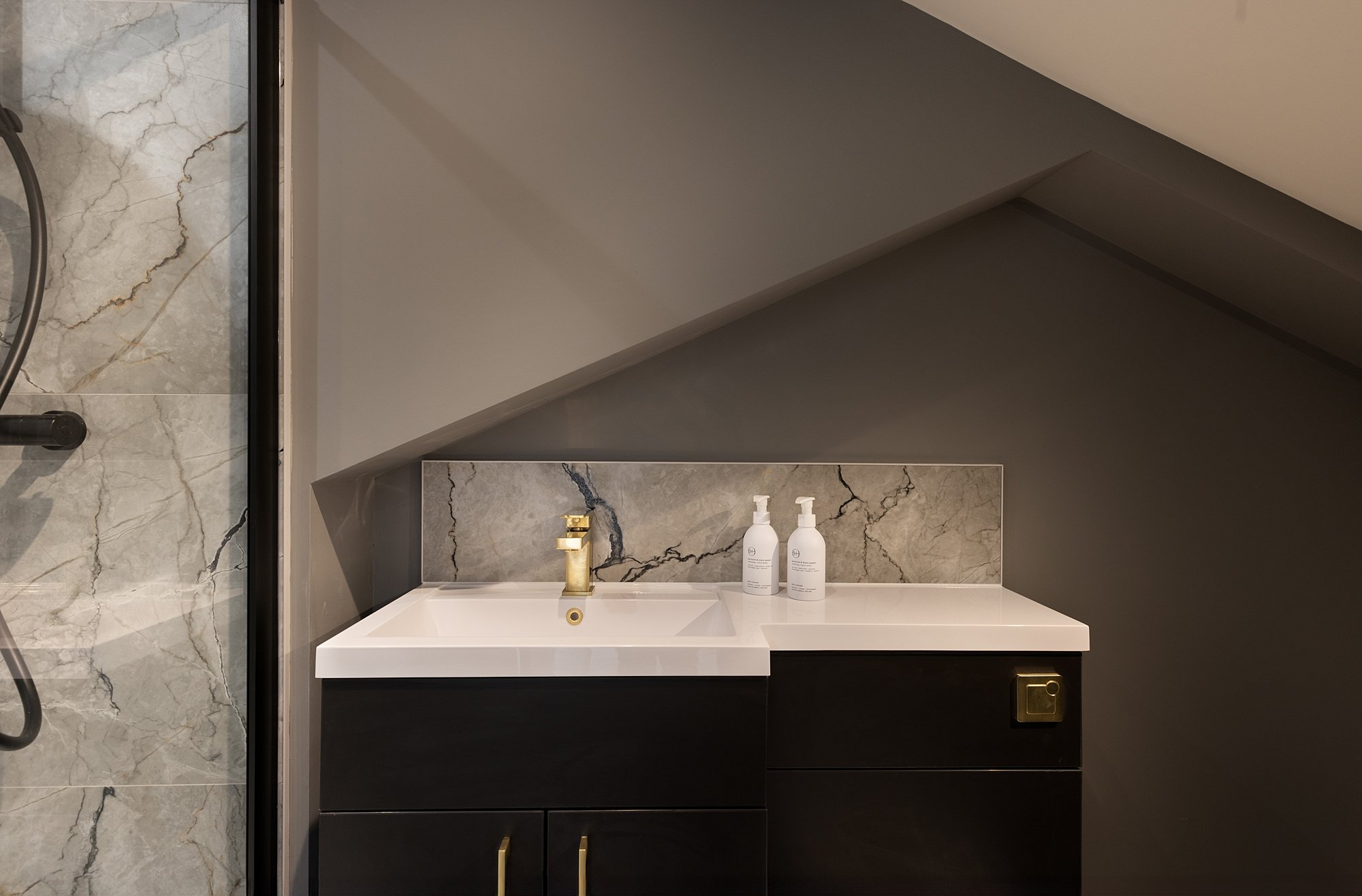 Bathroom vanity with black cabinet, white countertop, marble backsplash, and two bottles of soap or lotion, with part of a walk-in shower with marble tiles visible on the left.