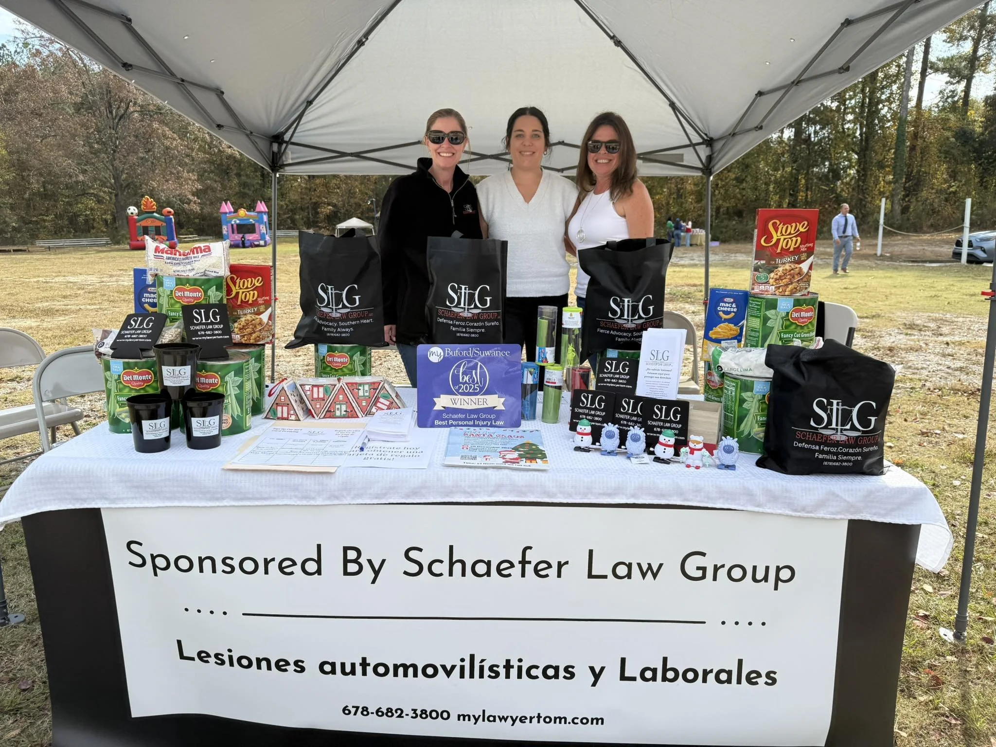 Three women smiling behind a table at an outdoor event. The table is covered with promotional items, bags, and a sign for Schafer Law Group. The background shows a grassy area with trees, a bounce house, and people walking nearby.