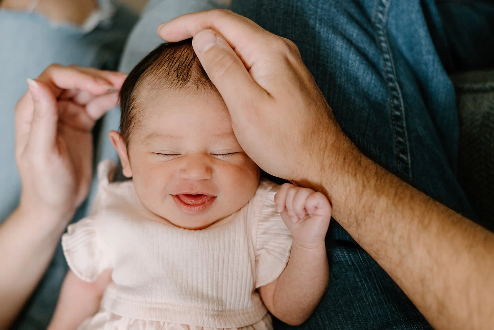 Close-up of a smiling newborn baby with closed eyes, lying on a person's lap, with one fist near their face and an adult's hand gently resting on their head.