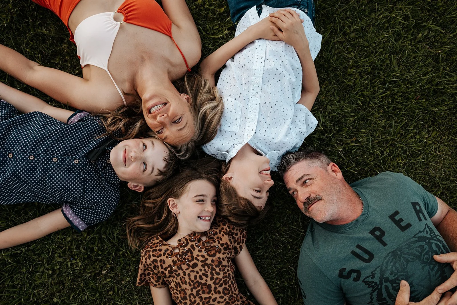 A family of two adults and three children lying on the grass and smiling at each other.