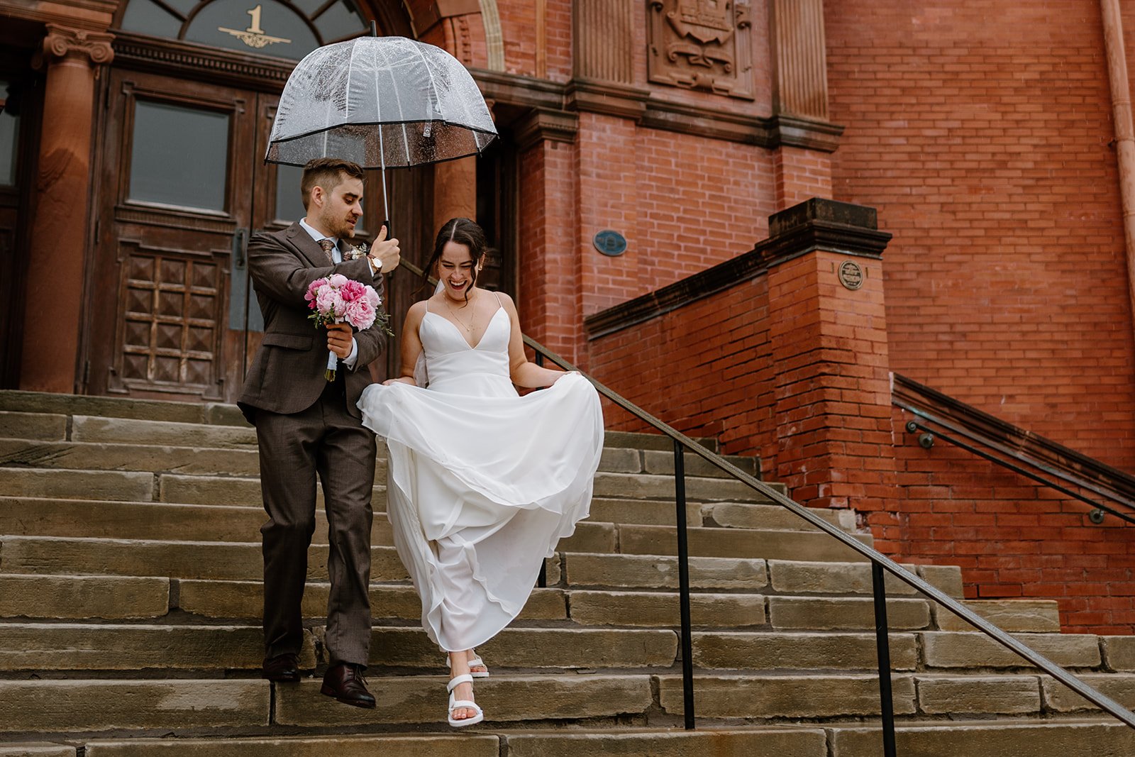 A newlywed couple walking down the steps of a red brick building in the rain. The groom, dressed in a brown suit, holds a transparent umbrella and a pink flower bouquet. The bride, wearing a white wedding dress, is joyfully lifting her dress as they descend the stairs.