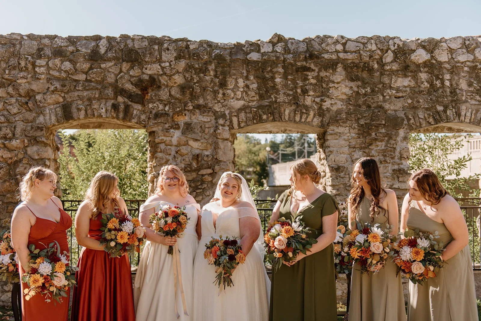 Eight women in wedding dresses and bridesmaids' dresses holding bouquets inside a stone archway during a wedding celebration.