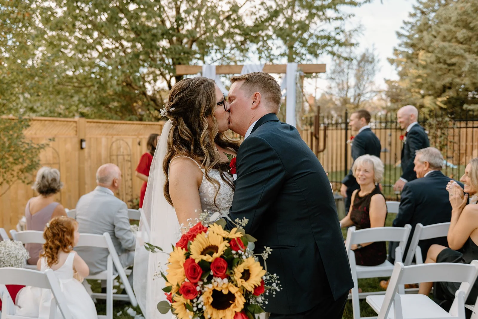 A newlywed couple sharing a kiss at their outdoor wedding ceremony, with family and friends seated around them and a wooden wedding arch in the background.