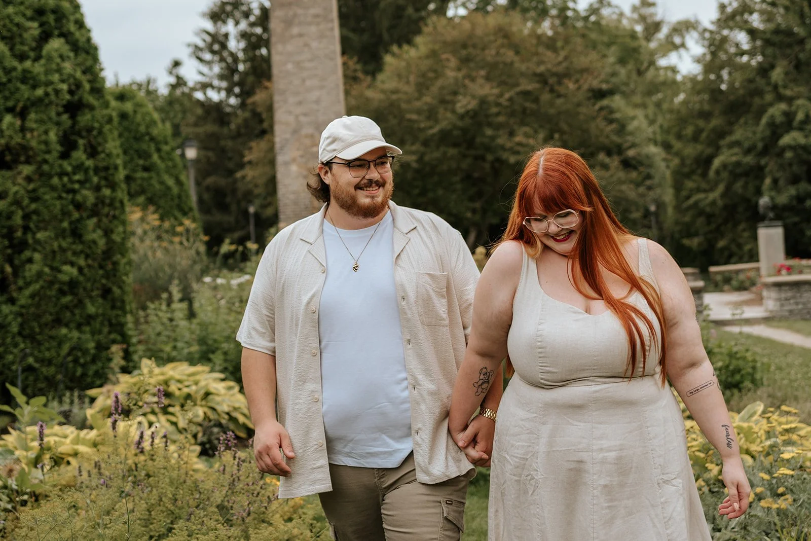 A smiling couple holding hands and walking through a garden with yellow flowers and green trees, with a stone tower in the background.