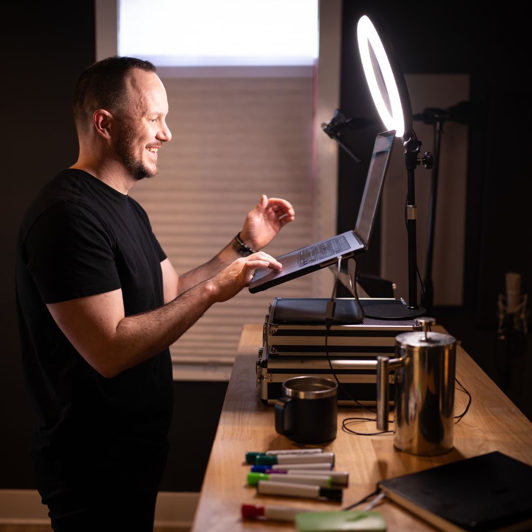A man smiling as he interacts with his laptop on a desk, illuminated by a ring light, with notebooks, markers, and a mug nearby in a well-lit room.