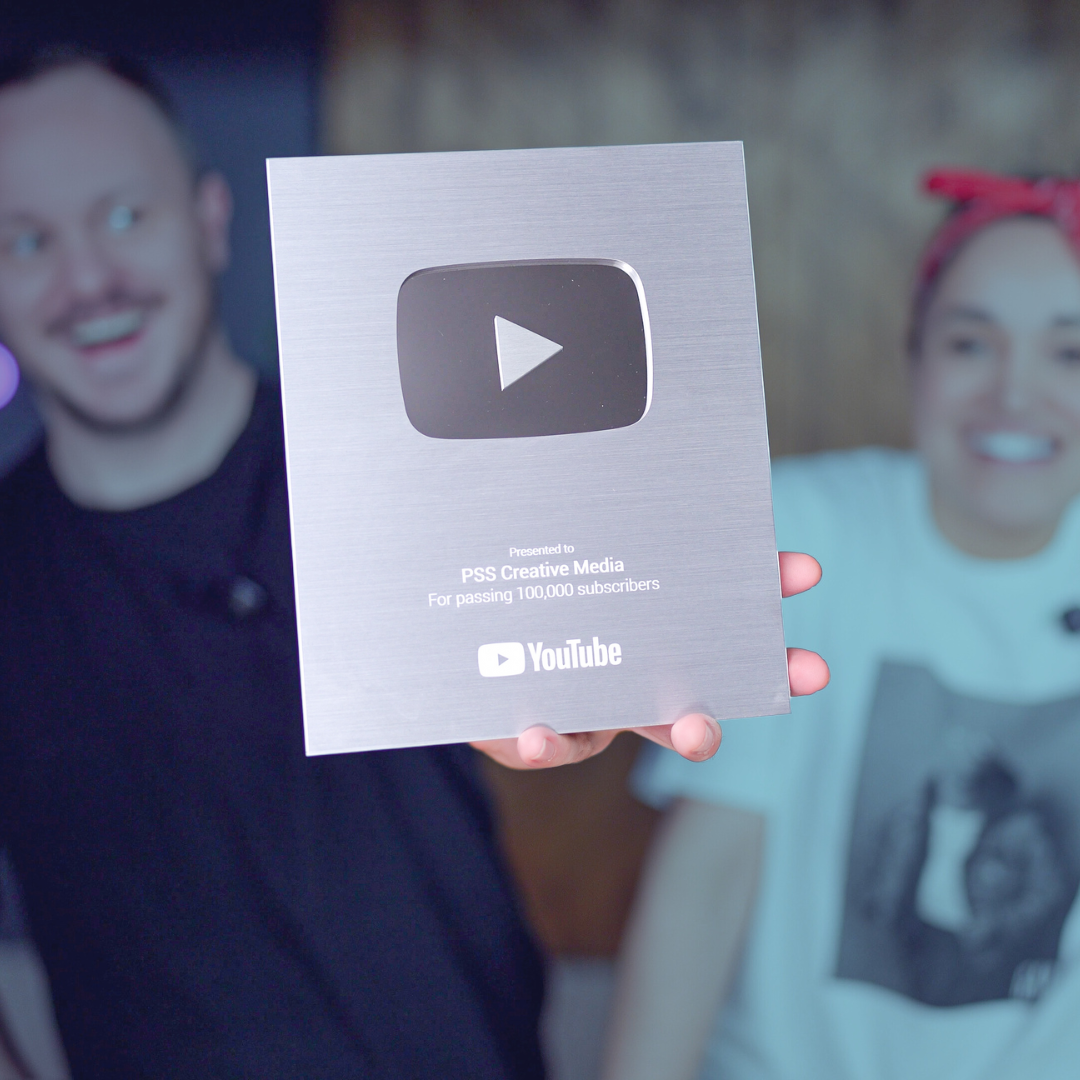 Person holding a silver YouTube silver play button award plaque for reaching 100,000 subscribers, with two smiling people in the background.