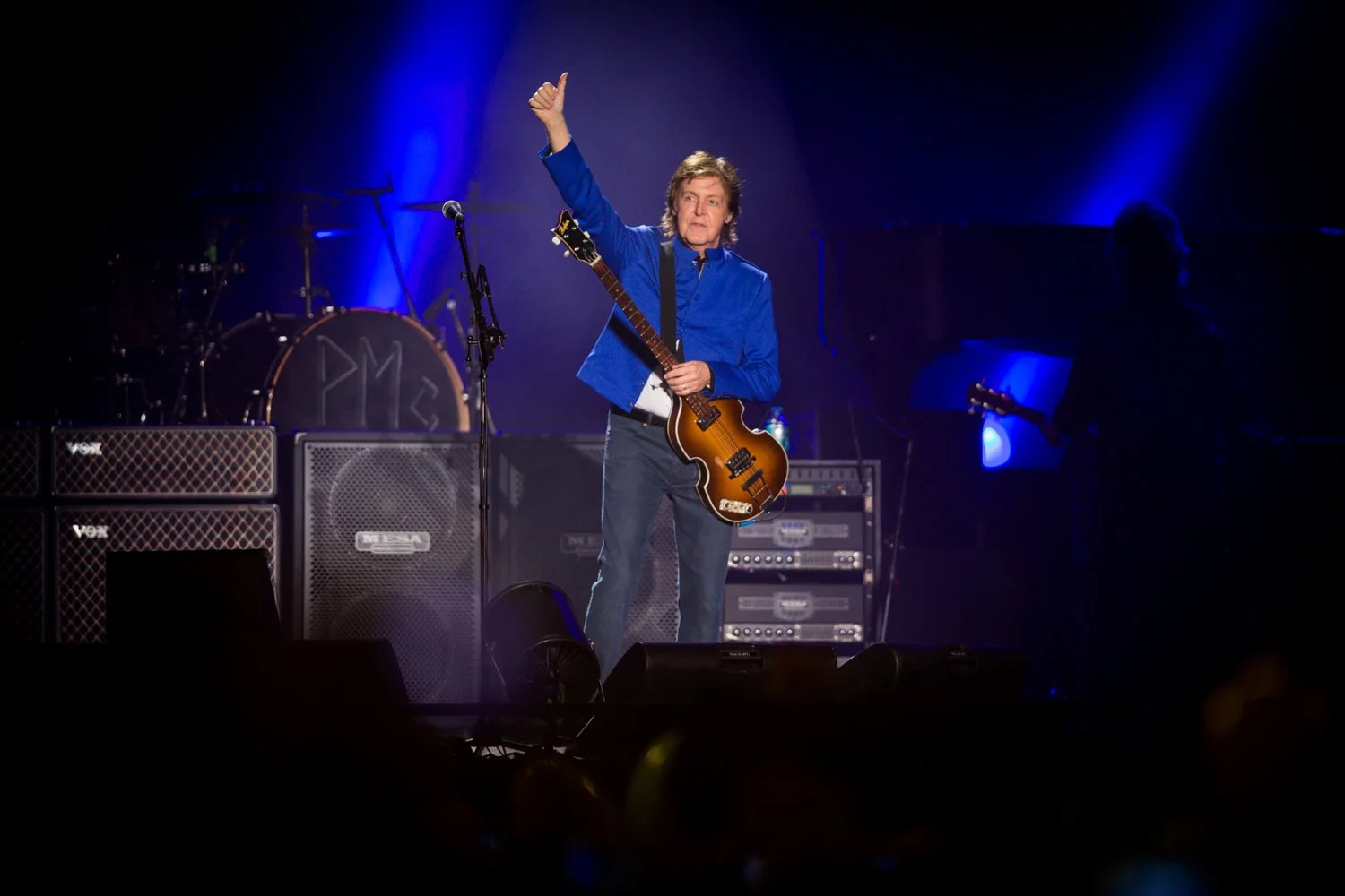 A musician on stage holding a guitar and giving a thumbs-up gesture, with stage lights and musical equipment in the background.
