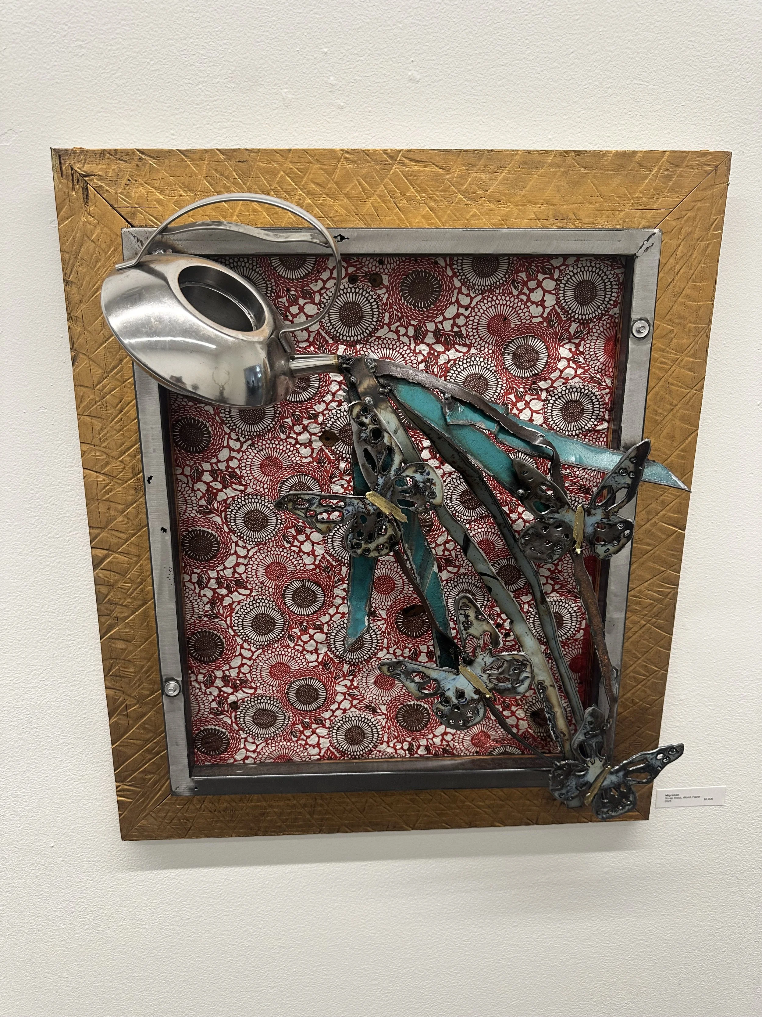 Mixed media art piece with metal butterflies and a watering can on a patterned red, white, and black background, framed in wood.