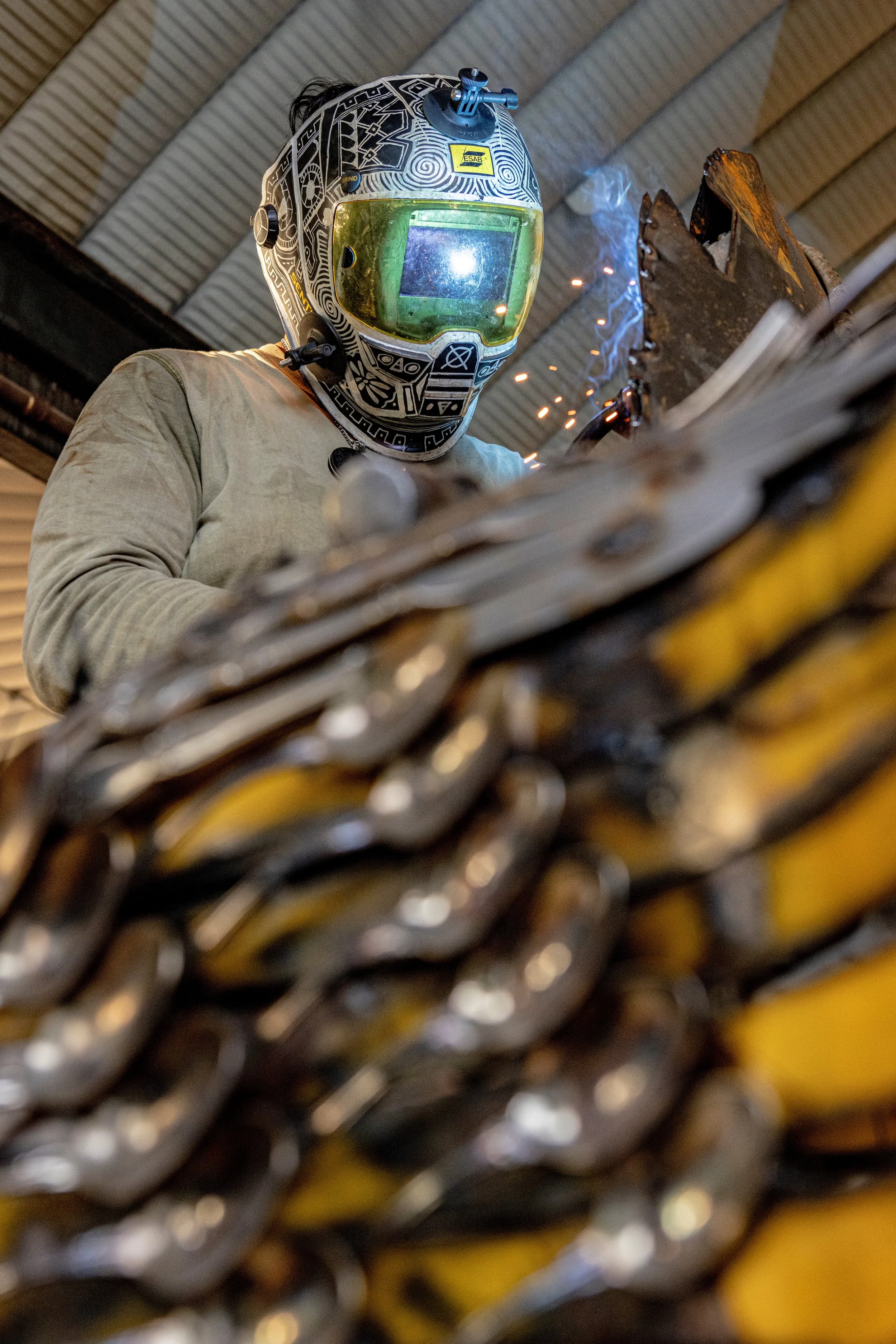 A worker welding metal chains, wearing a protective helmet with a yellow tint visor, indoors with a curved metal ceiling.