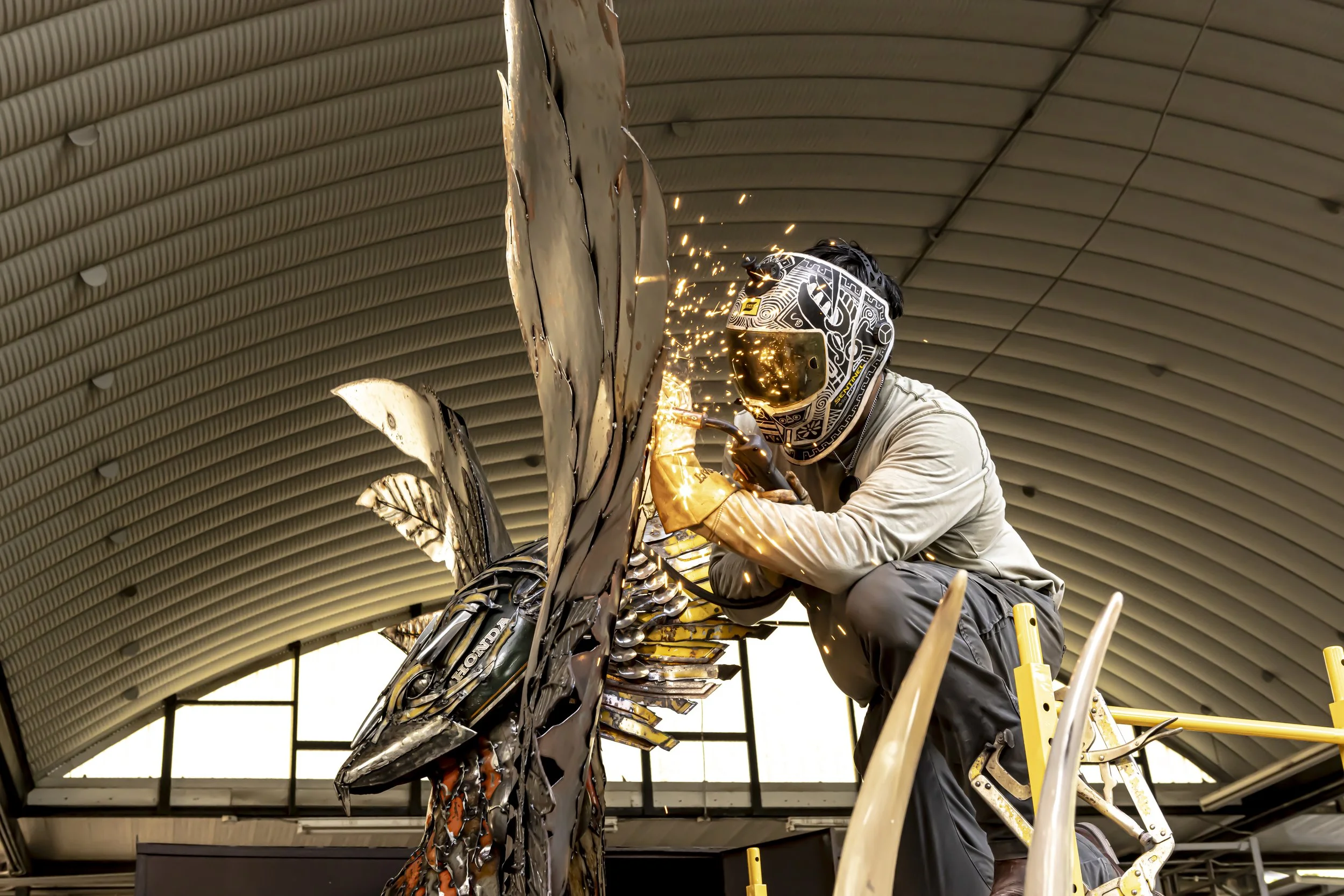 A person welding a large metal sculpture of a fish, wearing a helmet with a reflective visor, under an arched ceiling in an indoor workshop.