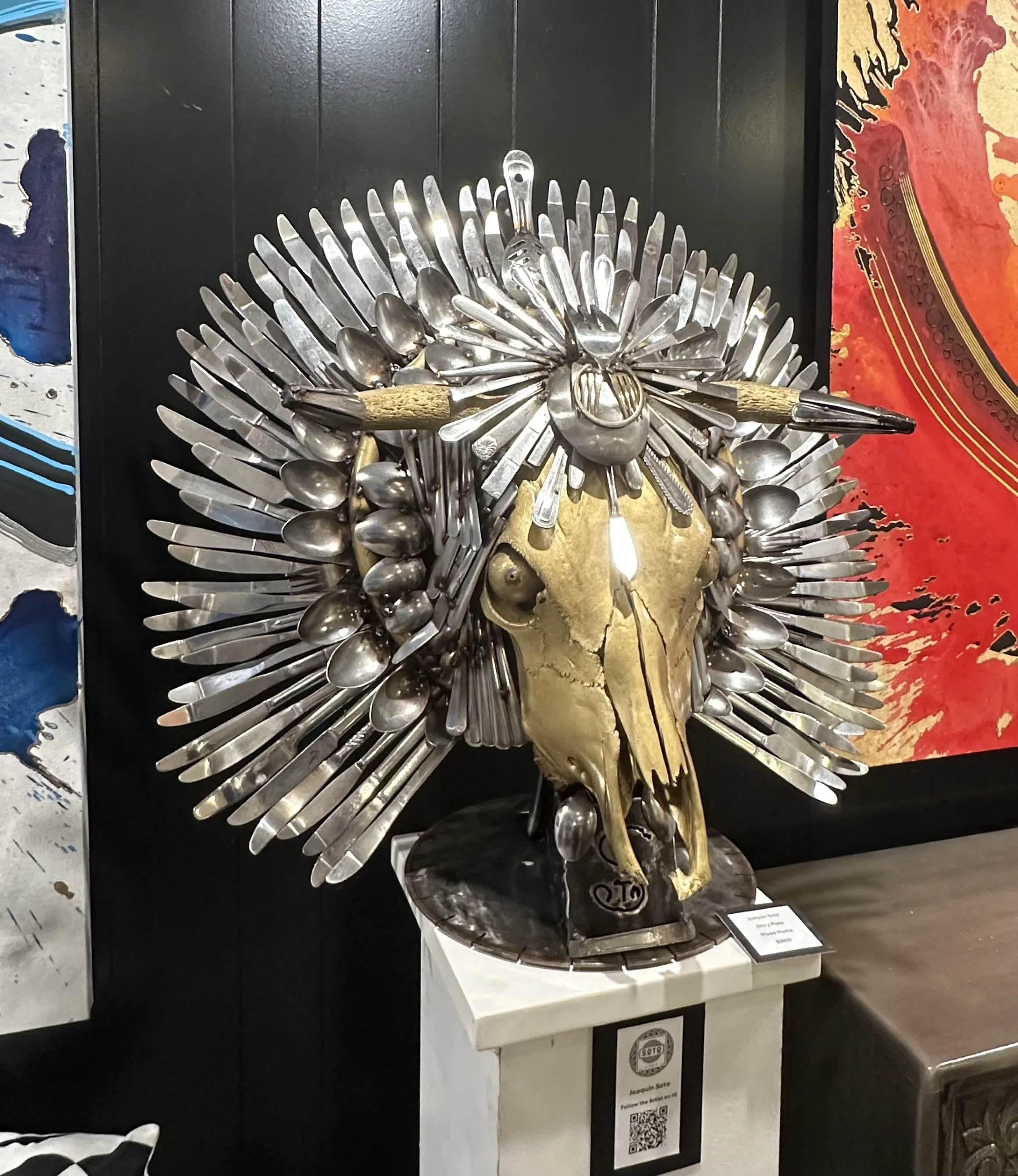 Art sculpture of a human skull decorated with spoons and utensils, with a gold-colored animal skull mounted on it, displayed on a white pedestal.