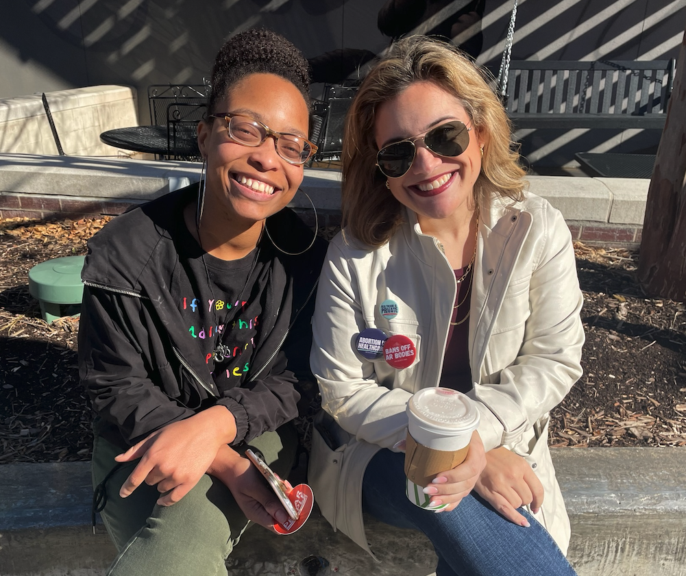 Two women sitting outdoors on a sunny day, smiling at the camera. One woman has curly hair, glasses, and is wearing a black jacket. The other has blonde hair, sunglasses, a white jacket, and multiple campaign pins, holding a coffee cup.