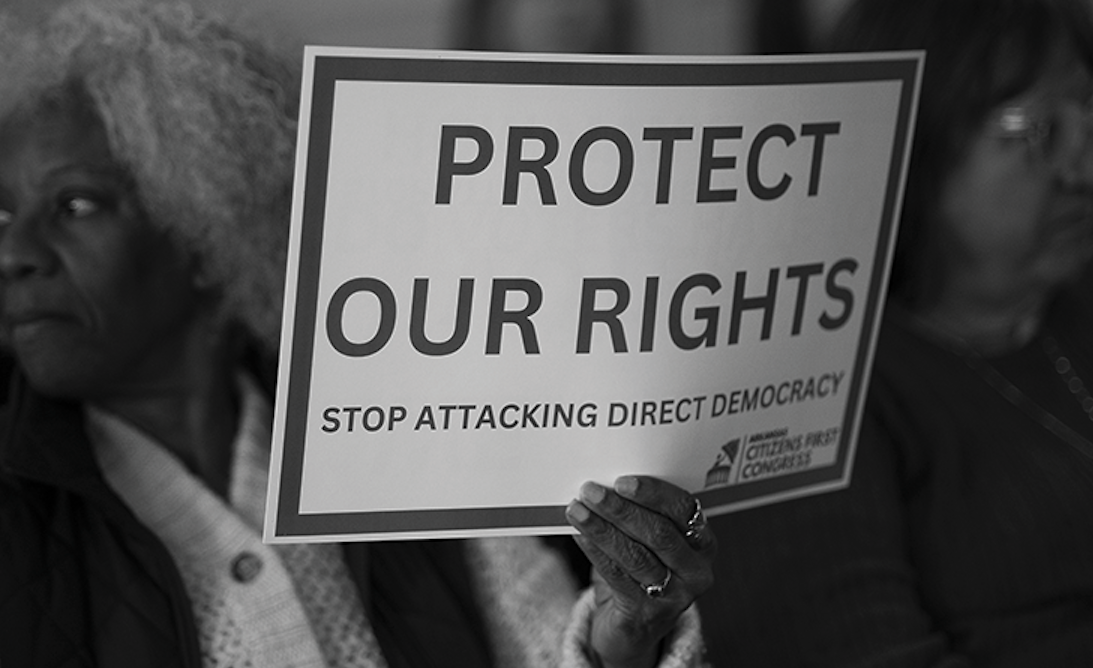 A person holding a sign that says "Protect Our Rights" and "Stop Attacking Direct Democracy," with two women visible in the background at what appears to be a protest or rally.