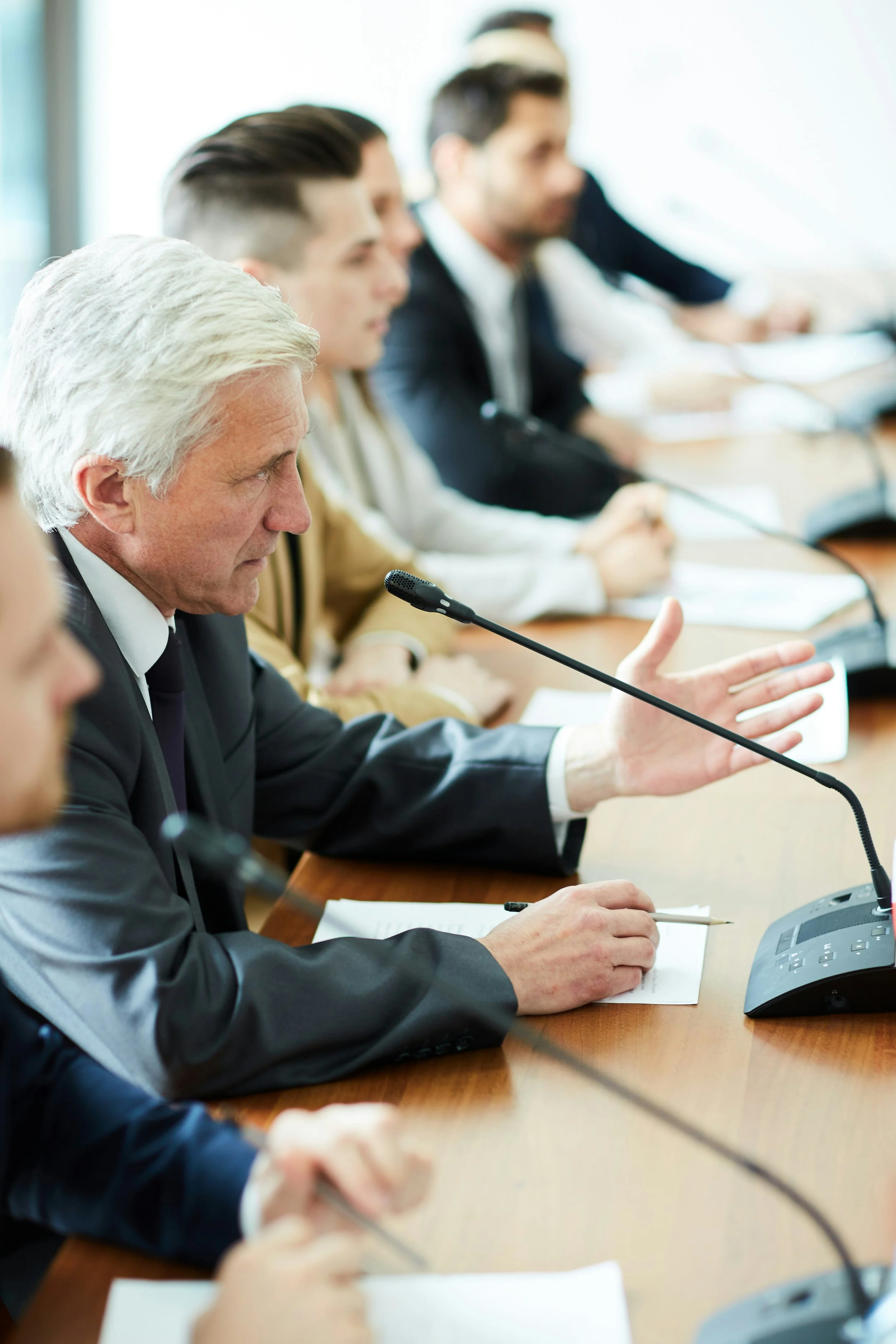 A business meeting with several people sitting at a conference table, engaged in discussion. An older man with gray hair in a suit is speaking and gesturing.