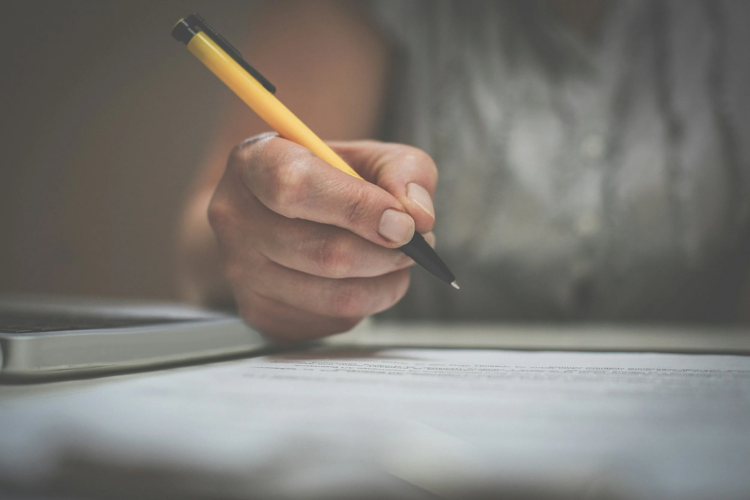 A person's hand holds a yellow and black pen, writing on paper on a desk.