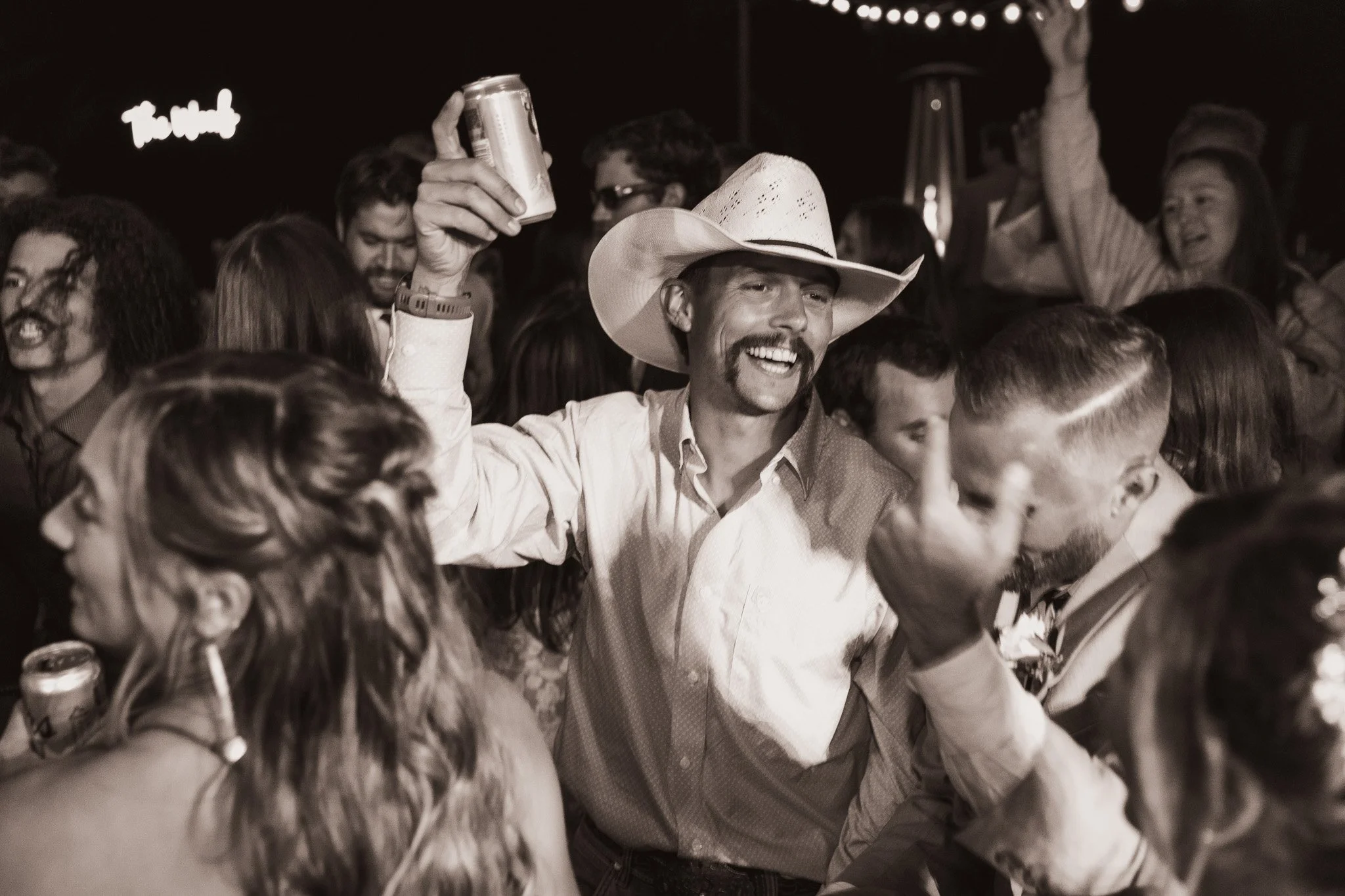 A group of people at a party, smiling, dancing, and enjoying drinks, with one man in the center wearing a cowboy hat.