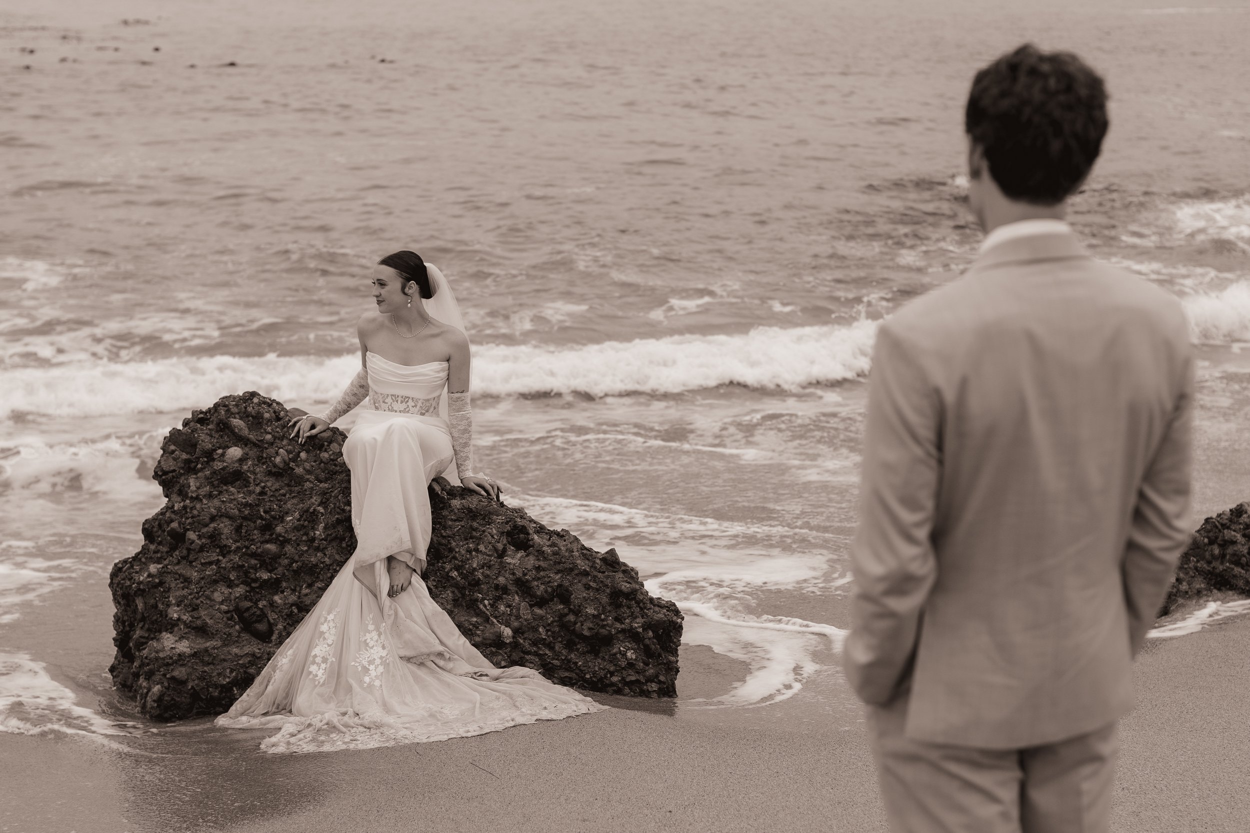 A woman in a wedding dress sitting on a rock at the beach, looking at a man in a suit who is standing nearby.