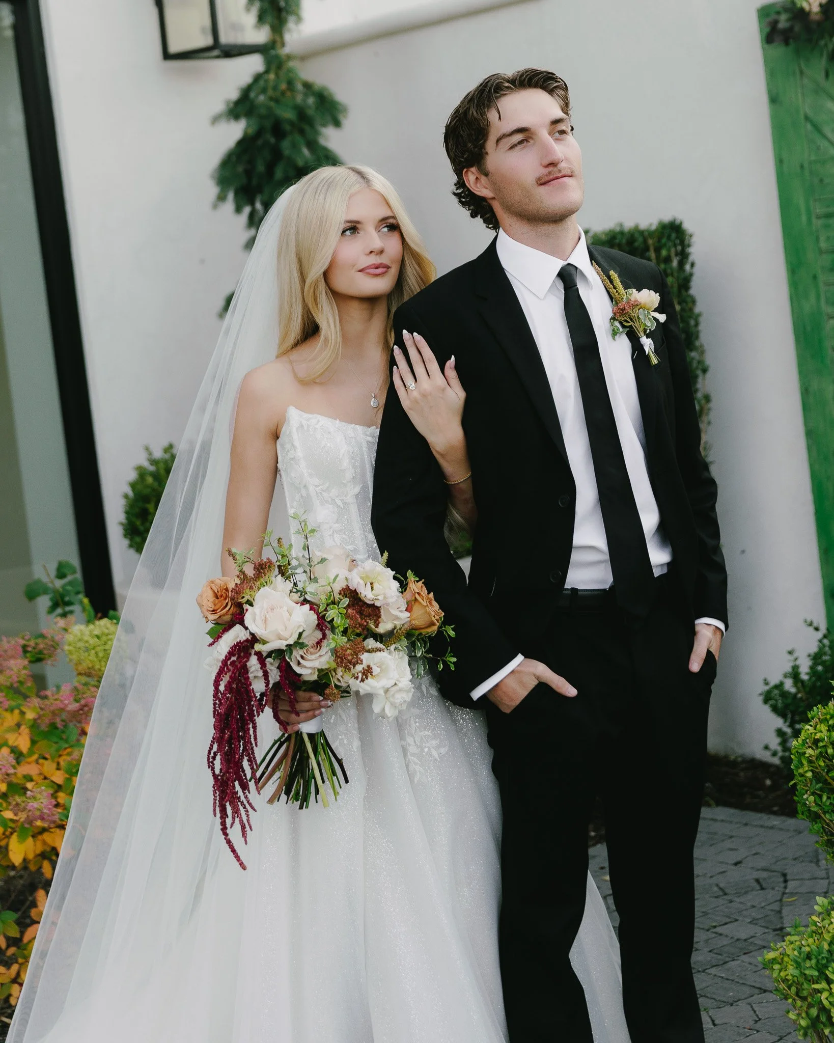 A bride and groom stand outdoors during a wedding ceremony. The bride has long blonde hair, wears a white wedding gown with lace details, a veil, and holds a large bouquet of white, pink, and burgundy flowers with greenery. The groom has brown hair, wears a black suit, white shirt, and black tie with a boutonniere. They are surrounded by greenery and flowers, with a white wall and a green door in the background.