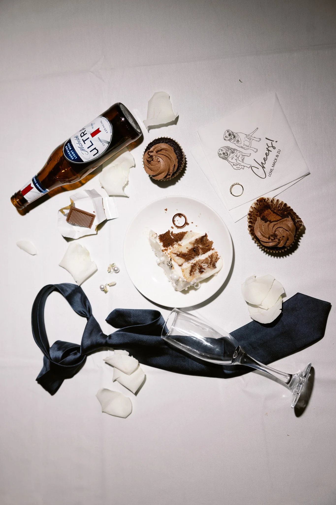 A partially eaten slice of vanilla cake with chocolate frosting, a Mr. right beer bottle, chocolate cupcakes, a piece of chocolate bar, a black ribbon, an empty wine glass, rose petals, pearl earrings, a card with a dog's drawing, and a ring on a whi