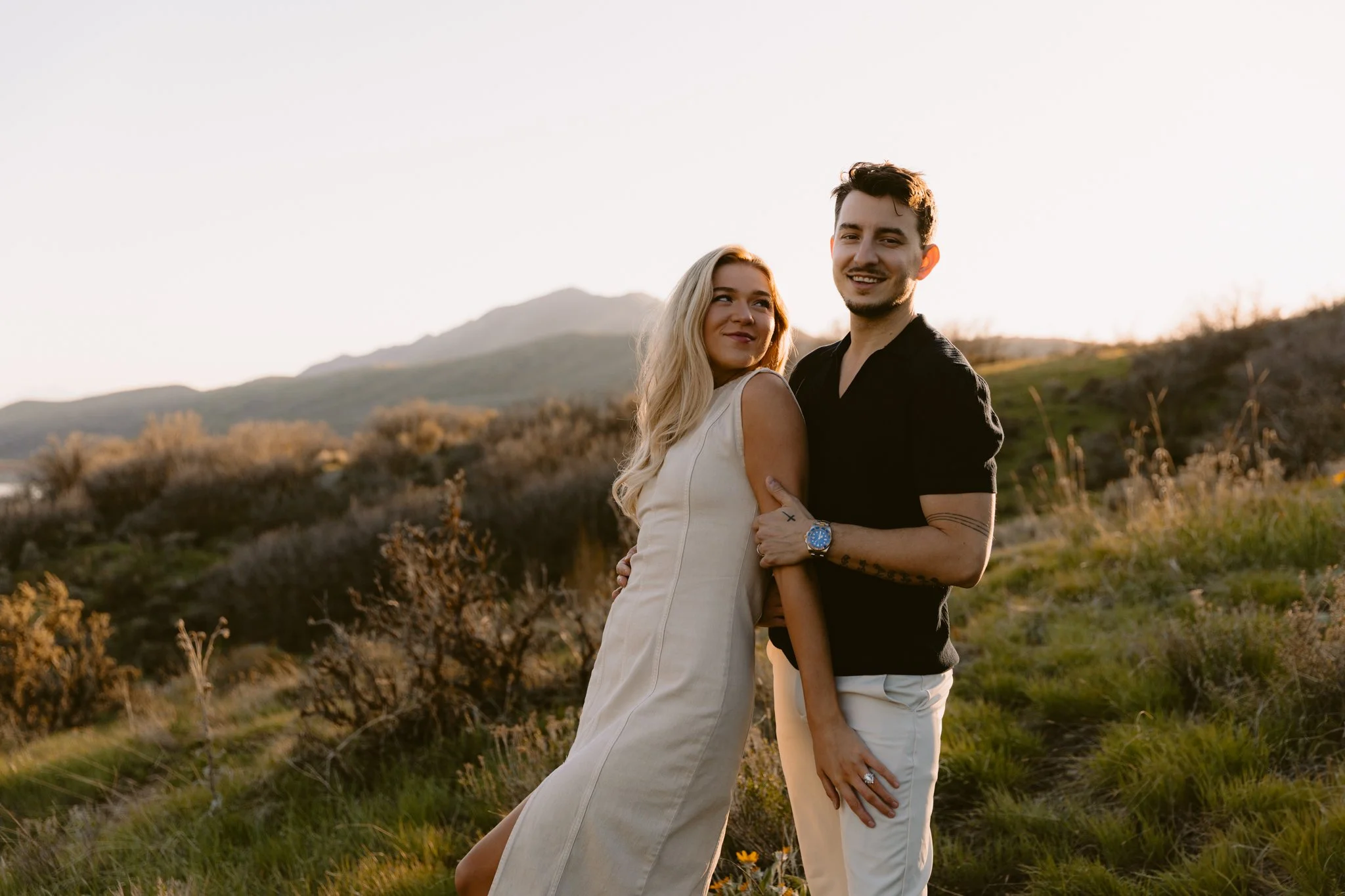 A couple standing outdoors in a grassy landscape with hills in the background during sunset, smiling and embracing each other.