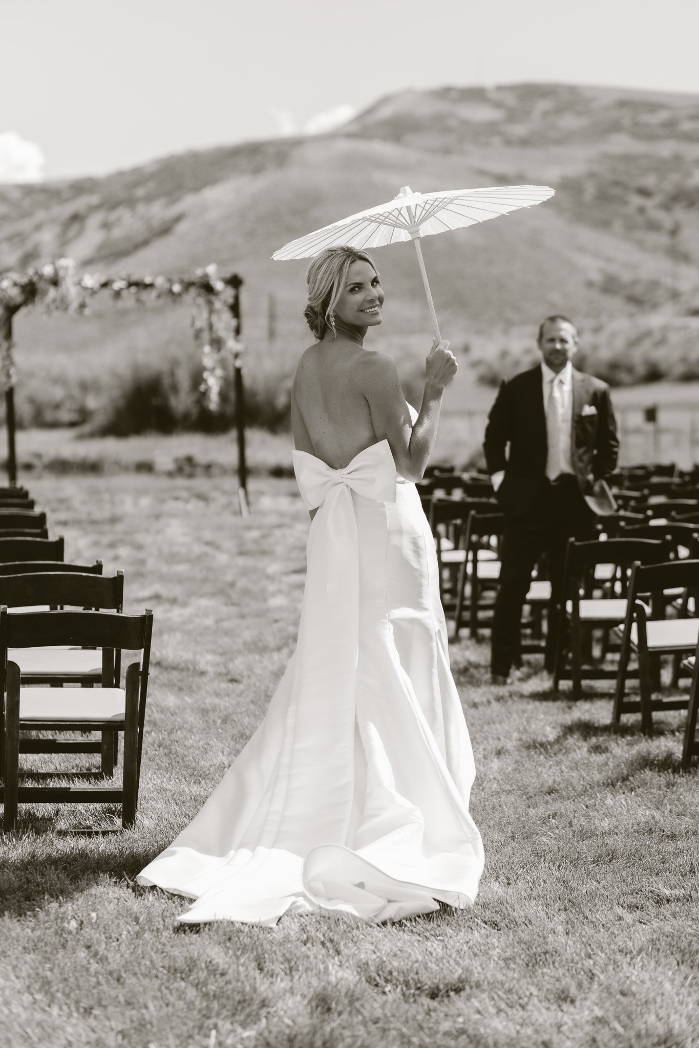 A joyful bride in a strapless wedding gown holding a parasol at an outdoor wedding venue, with chairs and a man in a suit in the background, mountain landscape