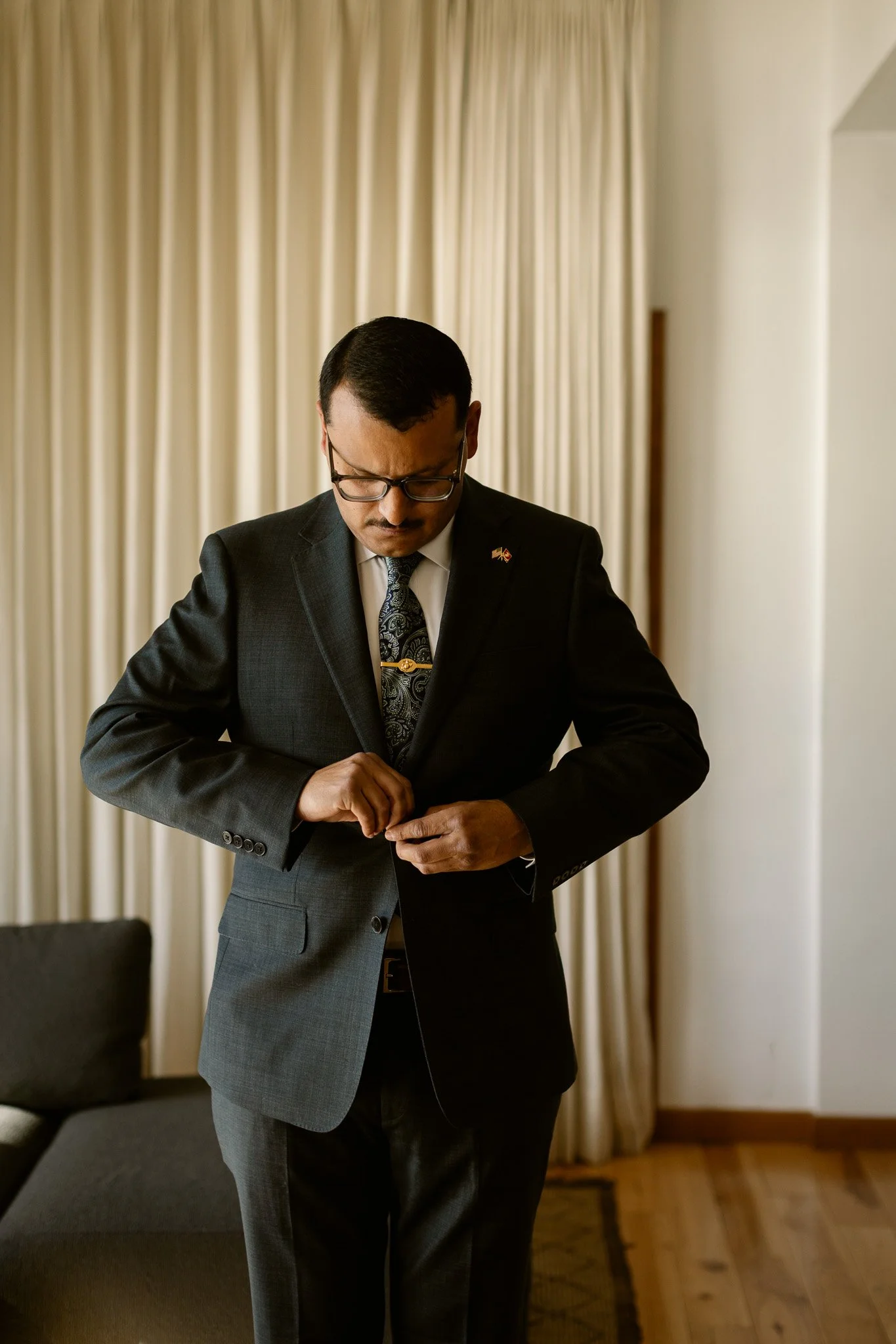 Man in a dark suit, patterned tie, and glasses buttoning his jacket in a room with beige curtains.