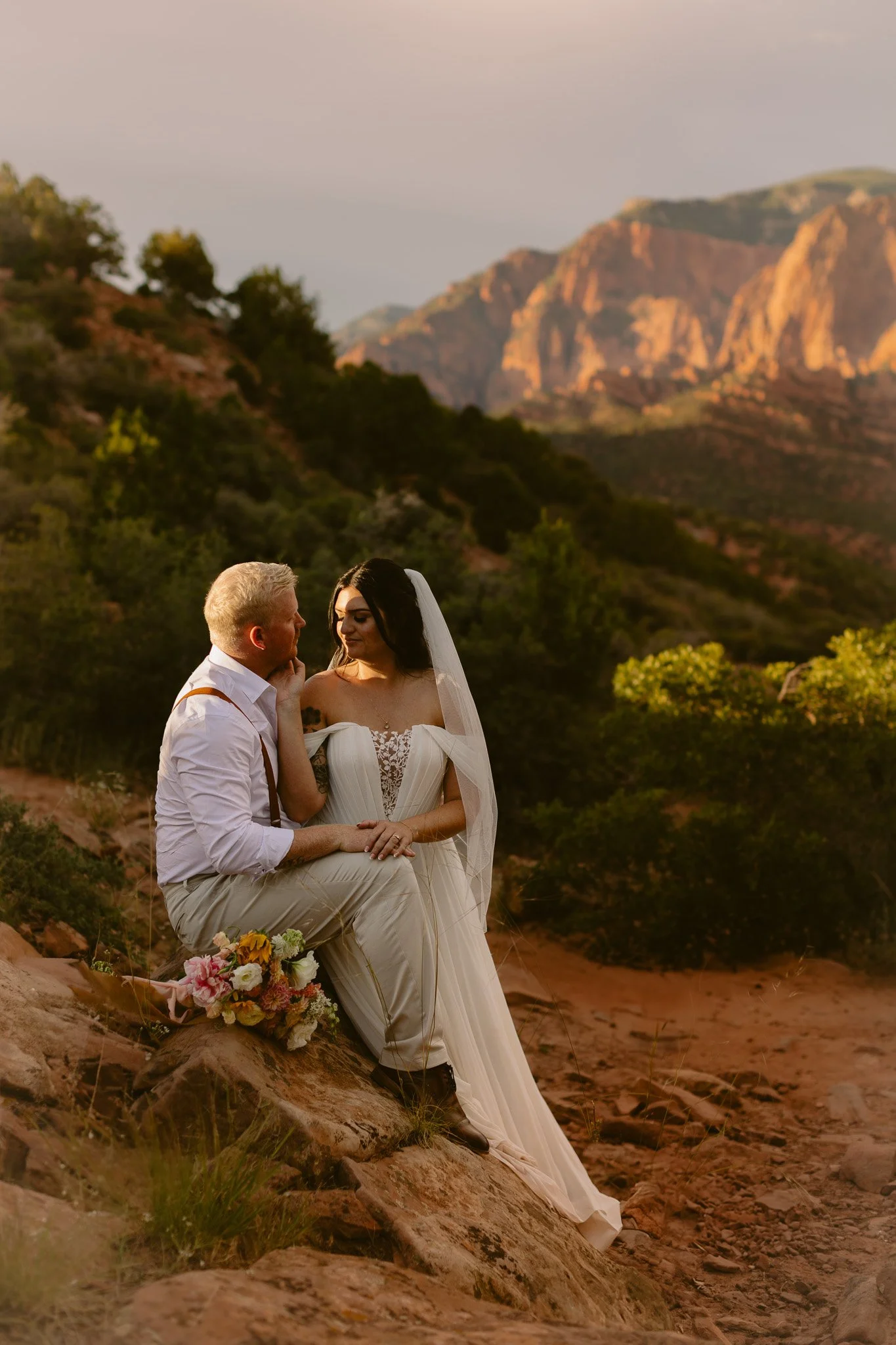 A bride and groom sitting on a rocky slope in a mountainous outdoor Zion setting during sunset, sharing an intimate moment, with a bouquet of flowers beside them.