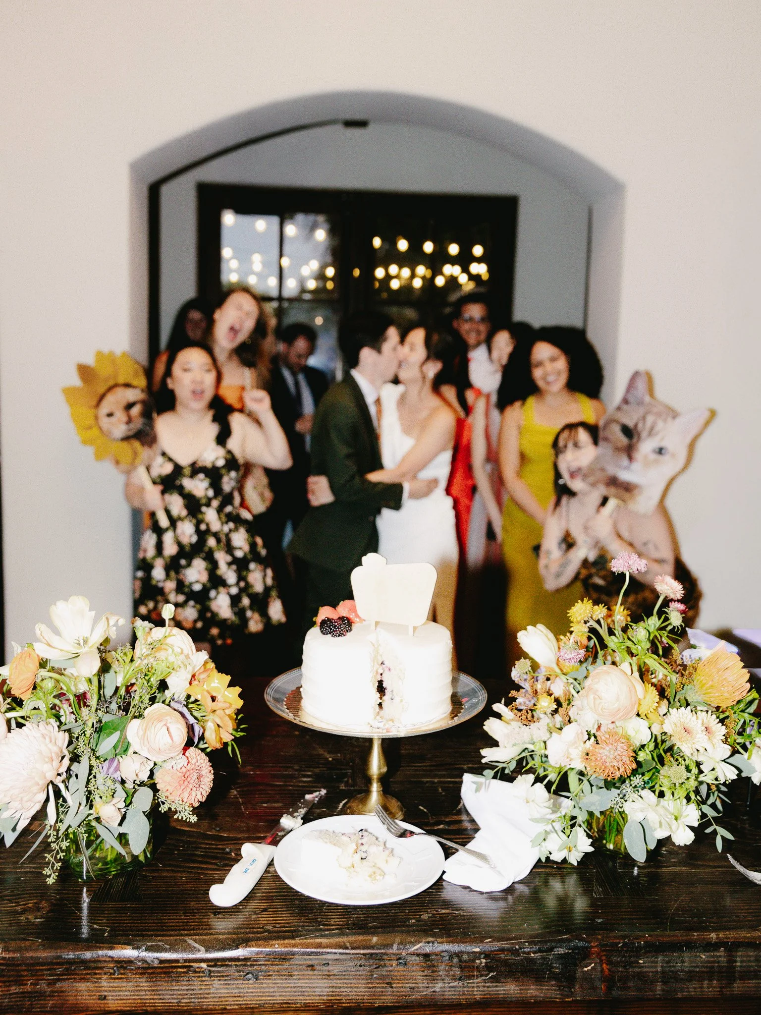 Group of people celebrating a wedding in front of a decorated cake, with floral arrangements on the table and partygoers making cheerful gestures.