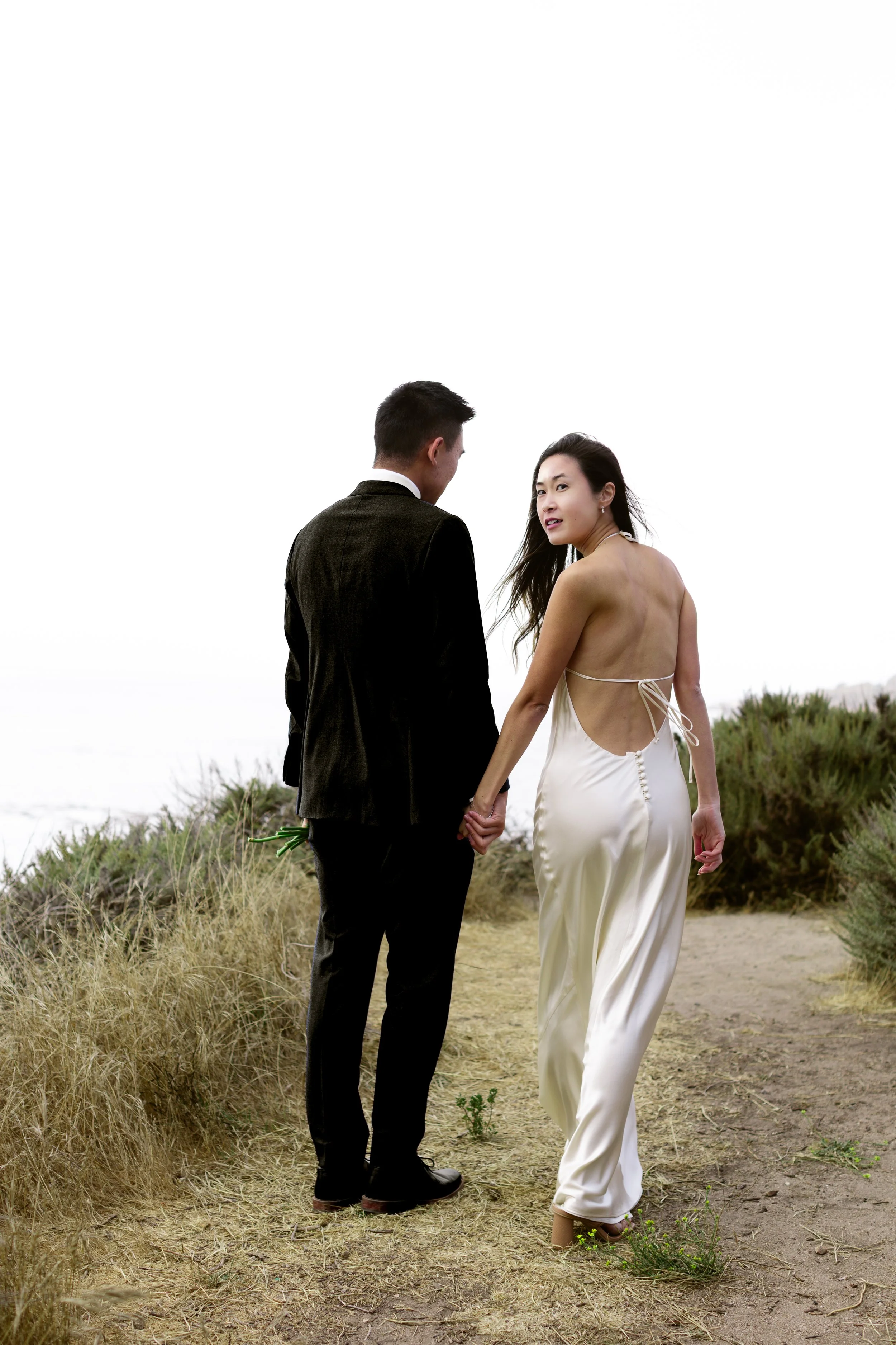 A couple dressed in wedding attire walking hand in hand on a dirt path surrounded by dry grass and bushes, with the ocean and cloudy sky in the background.