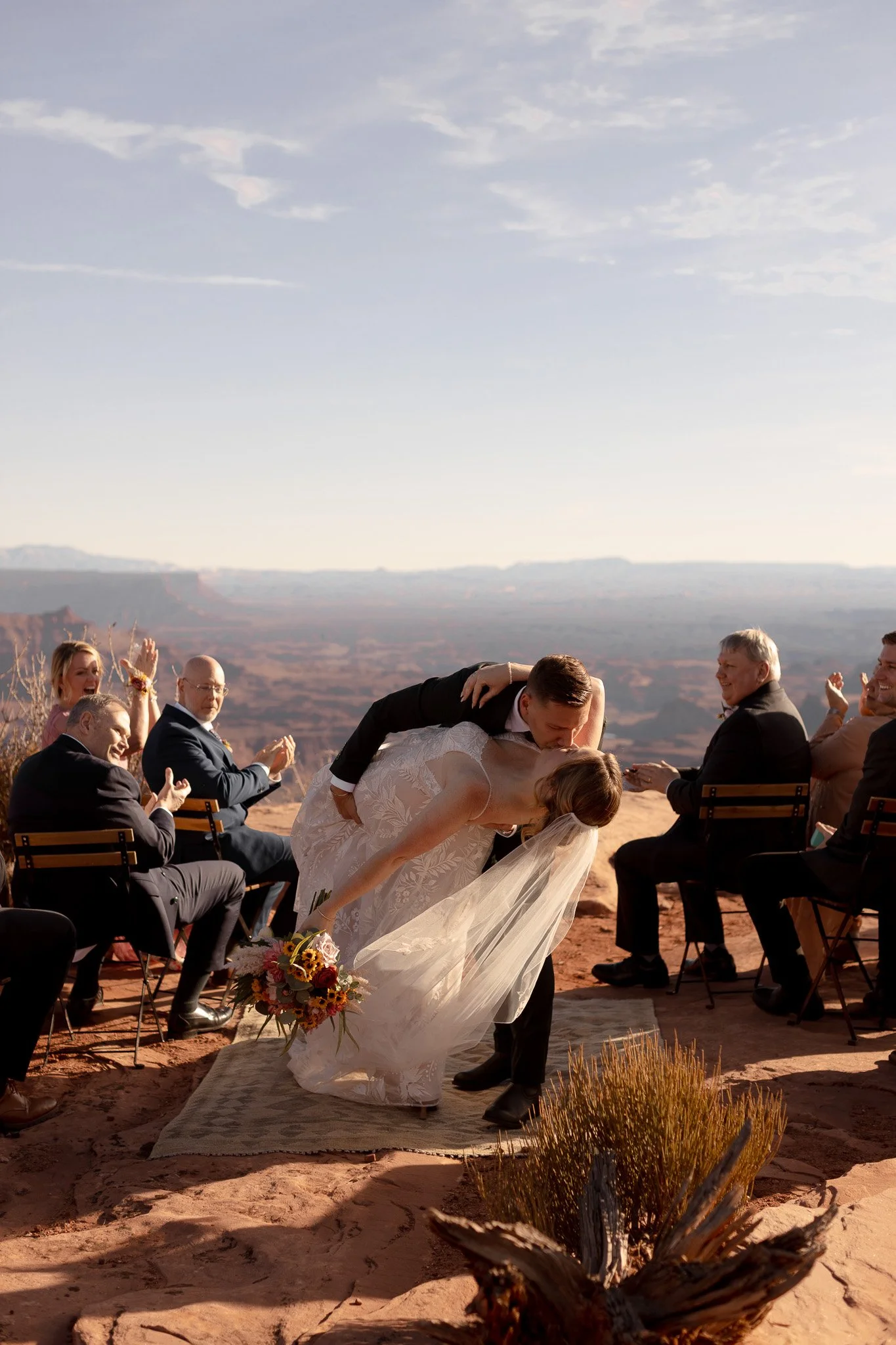 A bride and groom share a kiss during their outdoor wedding ceremony in a desert landscape, with guests seated on either side, some clapping and celebrating.