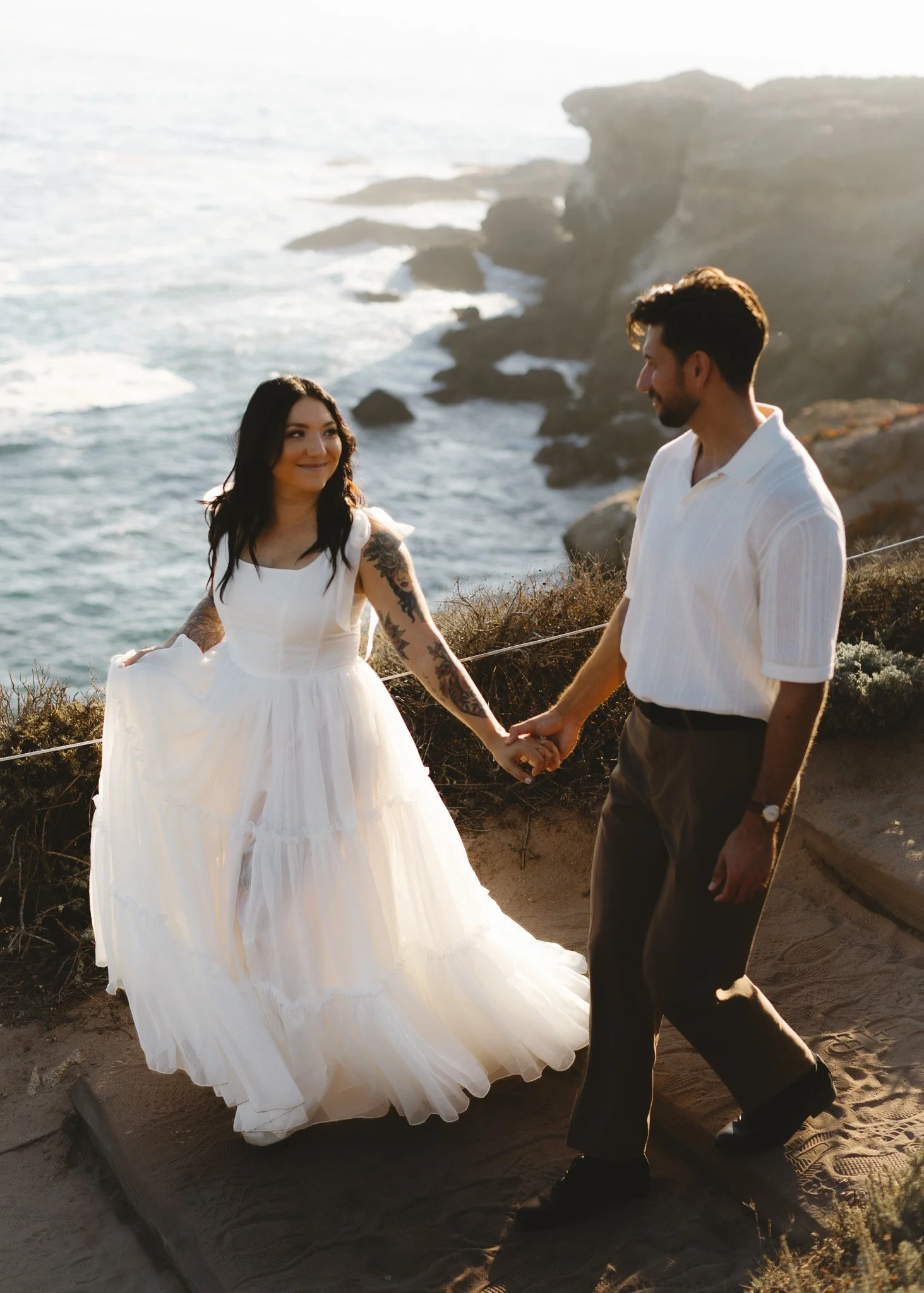 A couple holding hands on a sandy trail by the ocean at sunset, with cliffs in the background. The woman is wearing a white dress and the man is in a white shirt and dark pants.