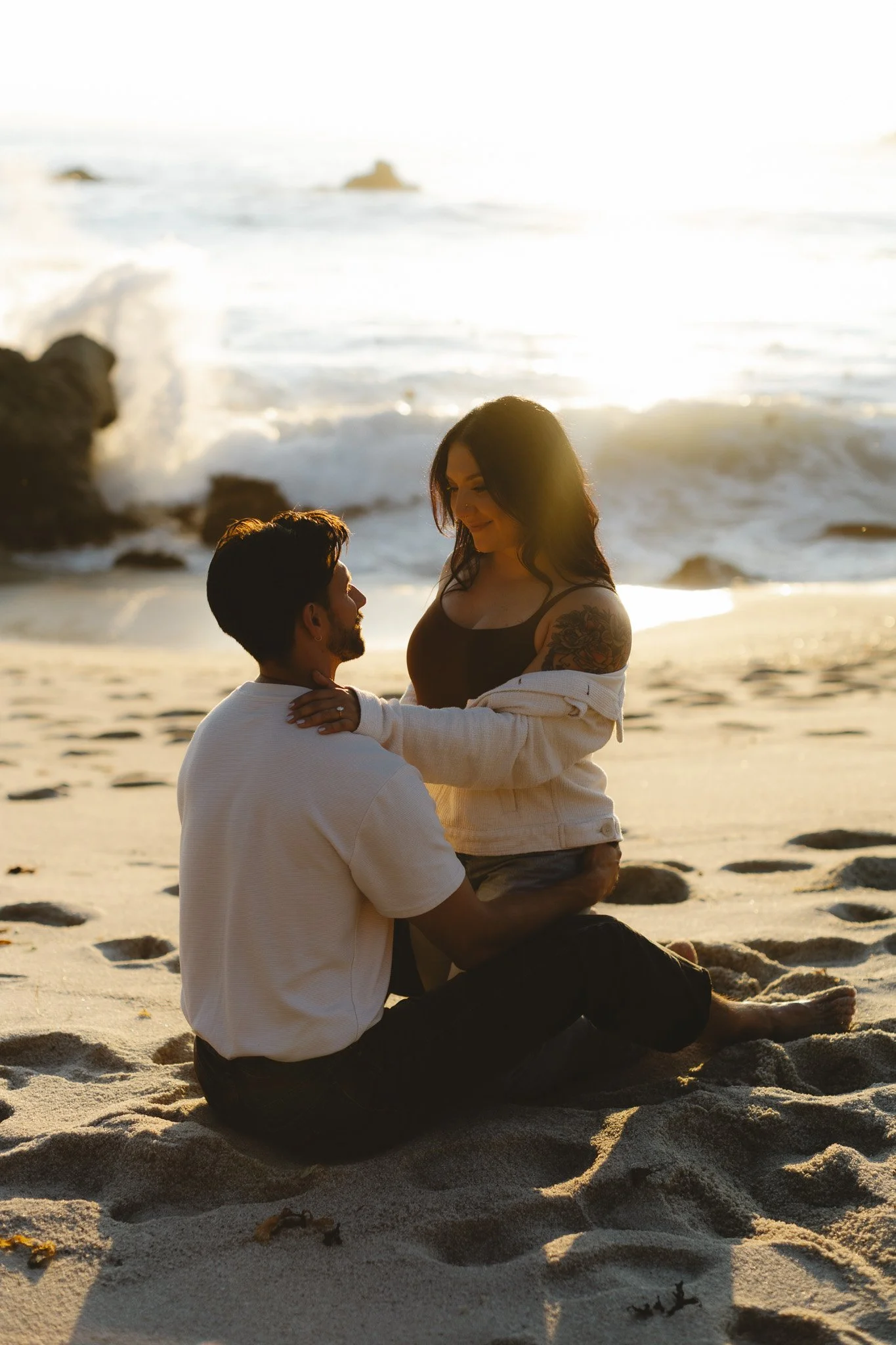 A couple sitting on the beach, facing each other, with the woman sitting on the man's lap during sunset, with waves in the background.