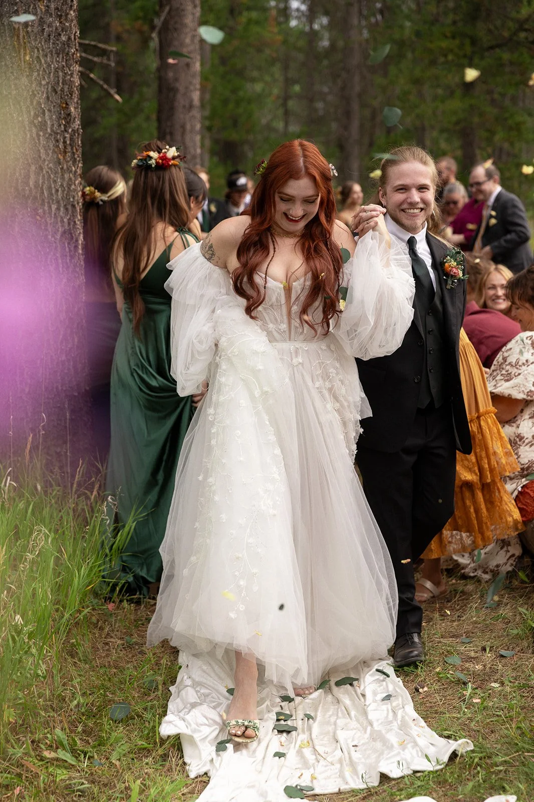Yellowstone wedding photographer captures bride and groom walking hand in hand post-ceremony as guests toss leaves and flowers near Swan Lake in Island Park, Idaho by Yellowstone National park.