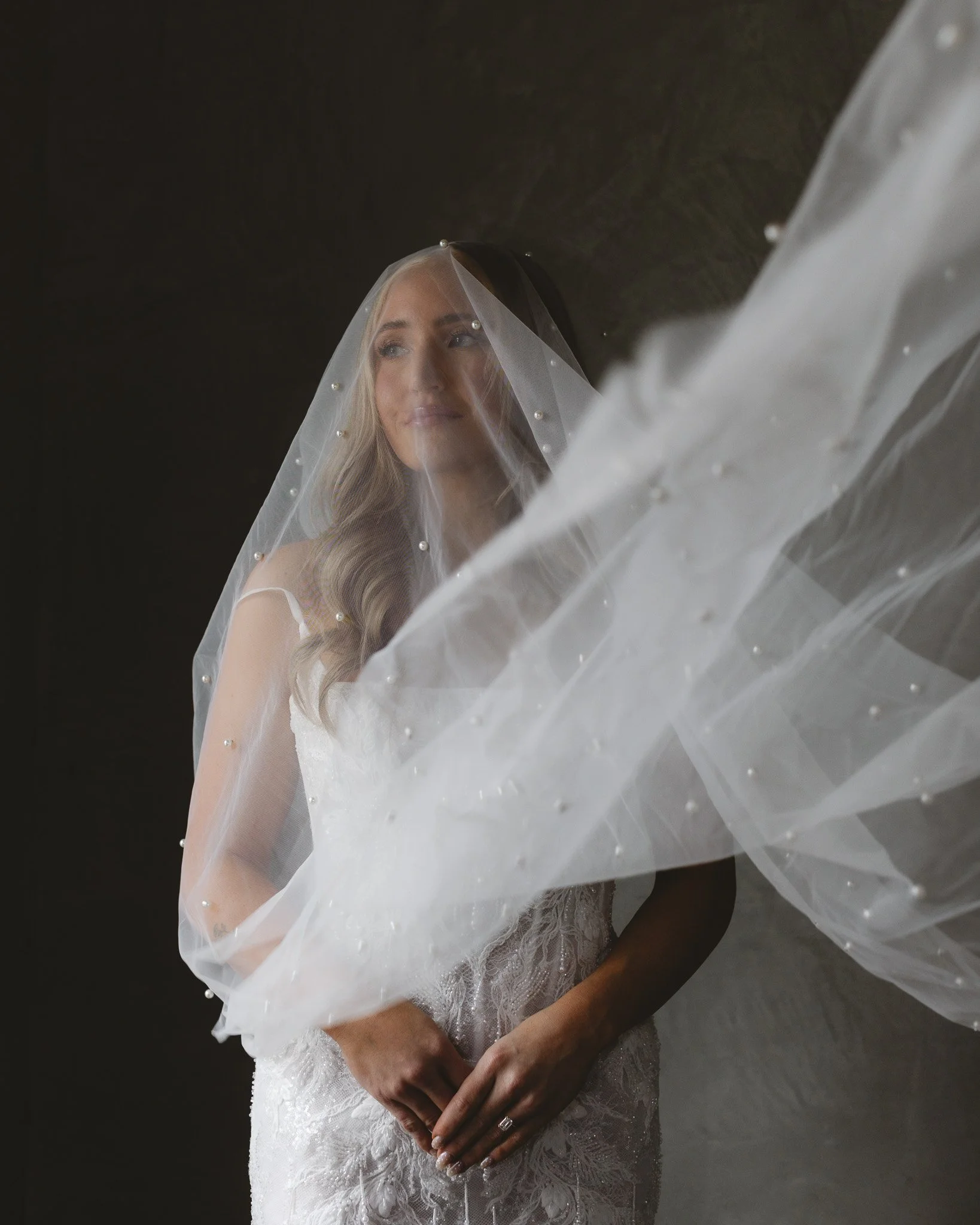 A bride in a wedding dress, looking thoughtfully with her hands clasped, wearing a veil decorated with small pearls, against a dark background.