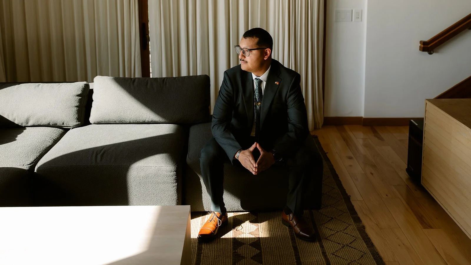 Man in a black suit sitting on a sofa in a sunlit living room.