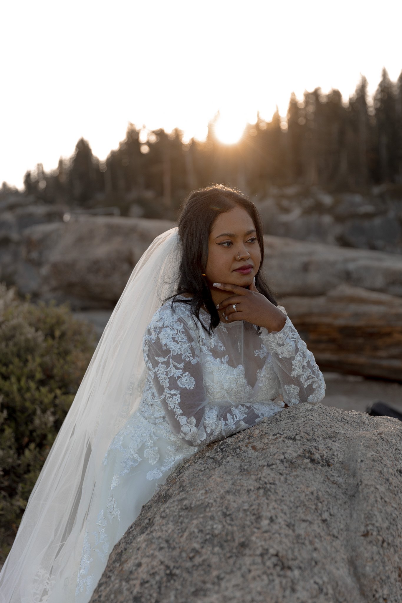 Bride in lace wedding dress with veil leaning on a rock, outdoor setting, sunlit trees in background.