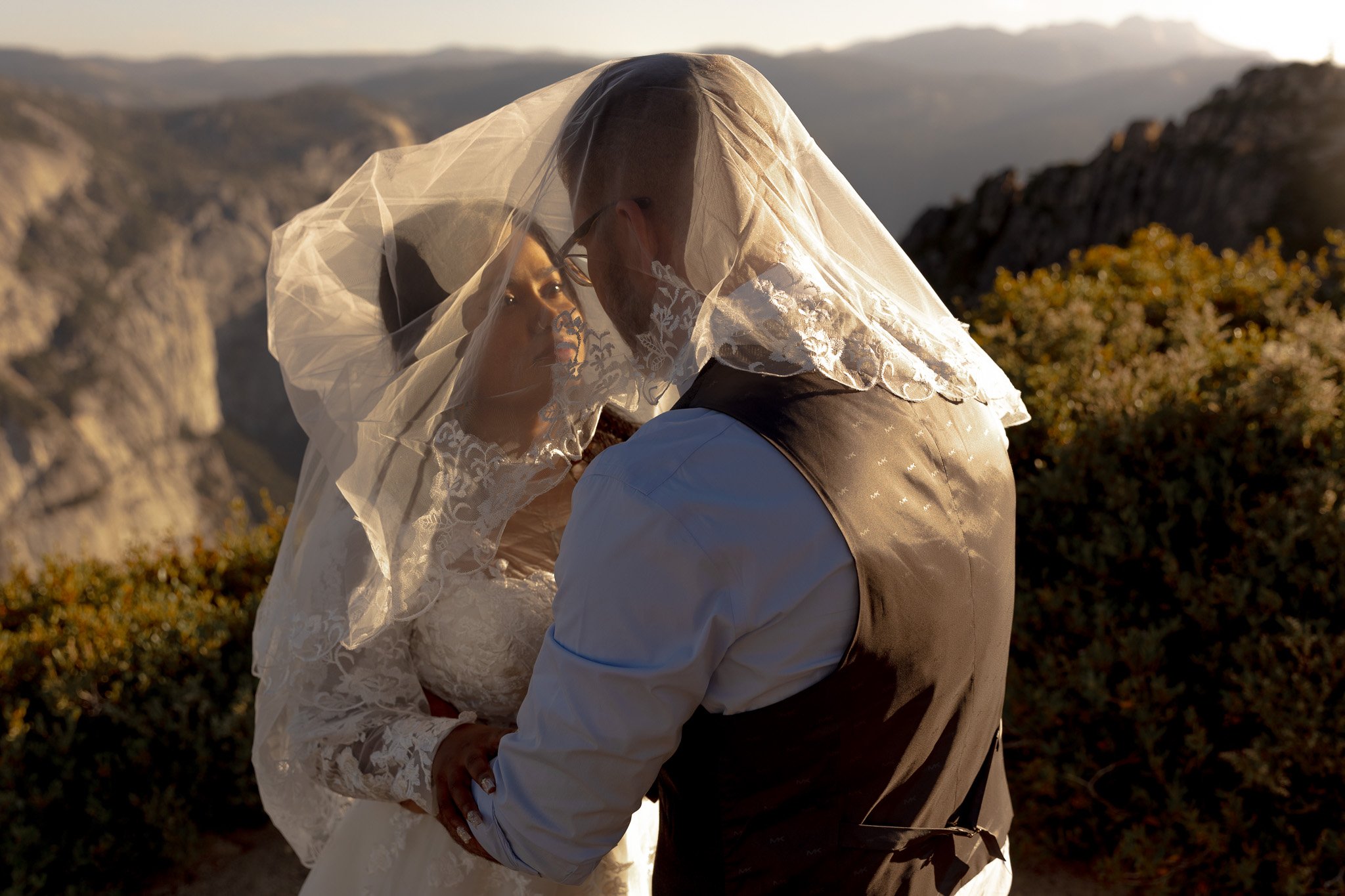 Bride and groom embracing under a veil in a mountainous landscape at sunset.