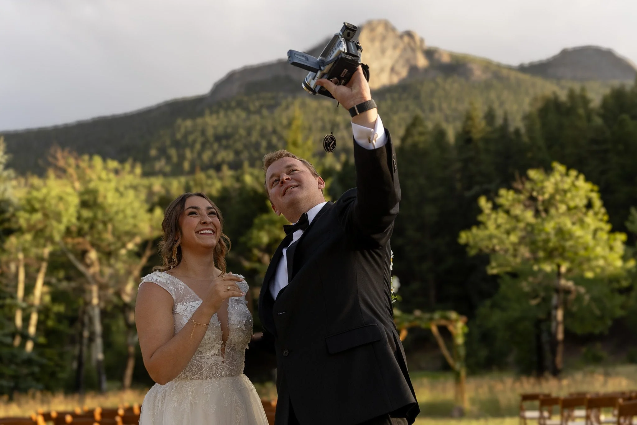 Bride and groom capturing a playful moment with a camcorder in Estes Park near Rocky Mountain National Park, surrounded by forest and peaks