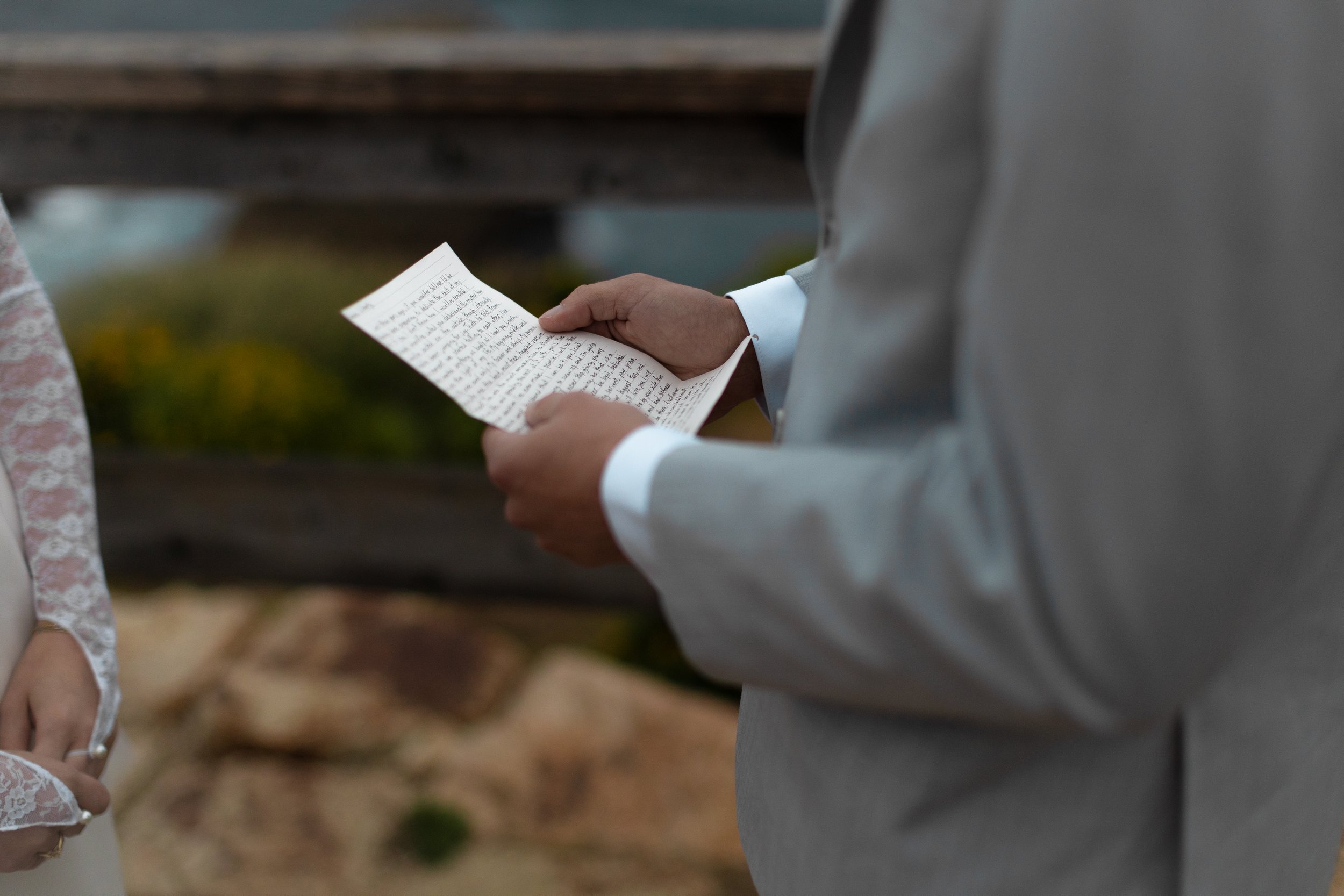A person in a gray suit holding a handwritten letter during a wedding ceremony. Another person in a lace dress is seen holding their hands near their waist.