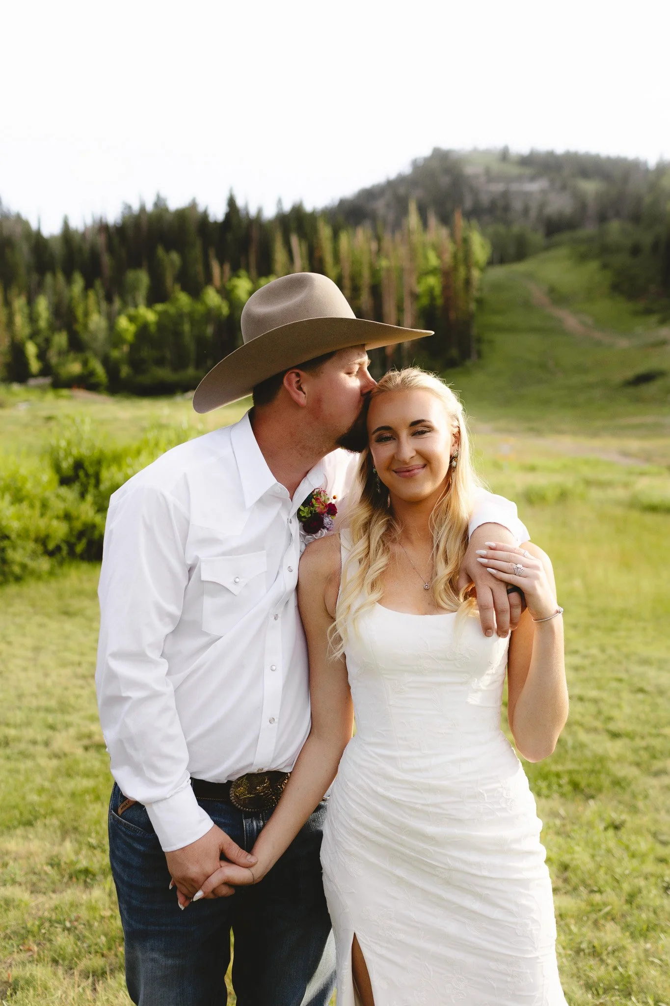 A man wearing a cowboy hat and a woman in a white dress stand together outdoors in a green landscape, with trees and a hill in the background. The man kisses the woman's forehead as they hold hands.