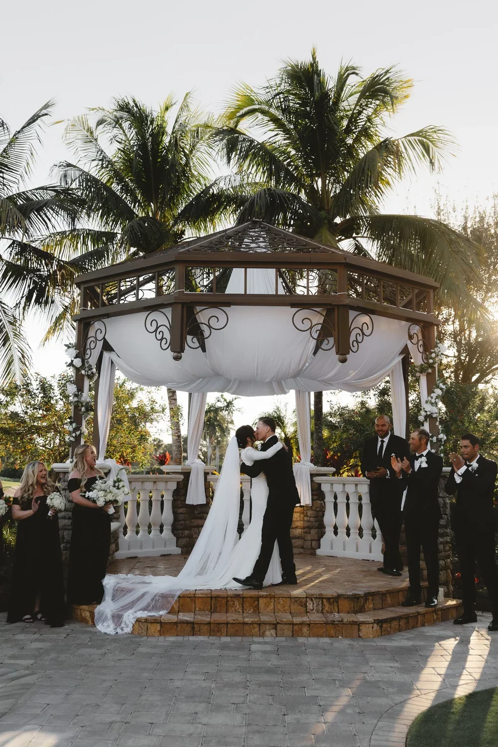 A couple is kissing at their wedding ceremony beneath a gazebo with white drapes and floral decorations, surrounded by bridesmaids and groomsmen, with palm trees in the background.