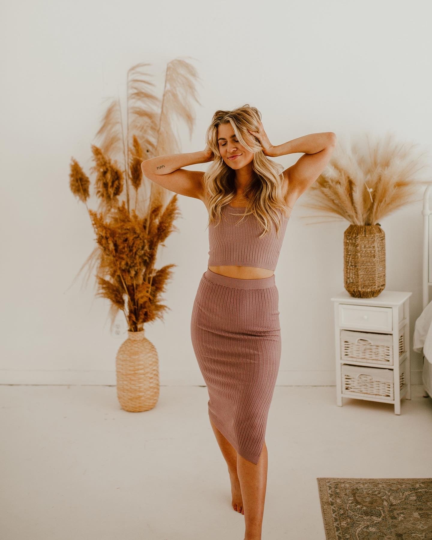 A woman in a matching mauve ribbed crop top and skirt stands in a room with neutral decor, with her eyes closed and hands in her hair, surrounded by large wicker vases filled with pampas grass.