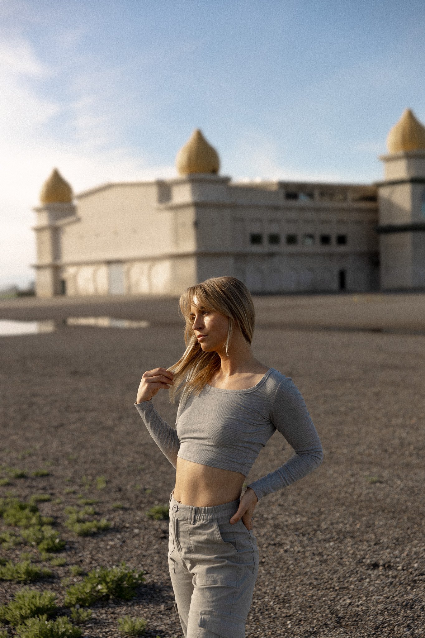 A woman in a gray crop top and beige pants standing outdoors near a large historic building with domed towers, under a clear sky.