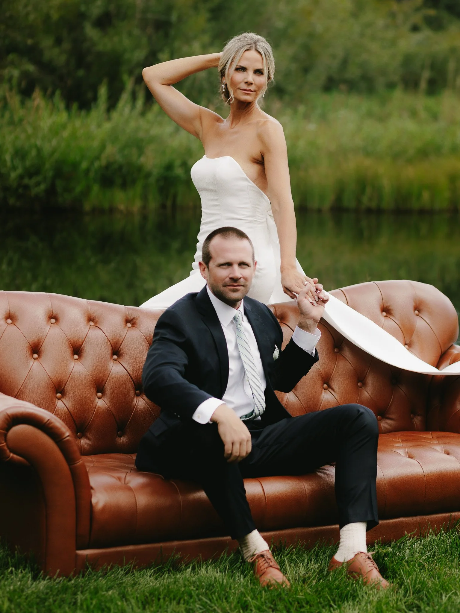 A woman in a white dress standing behind a seated man in a black suit on a brown leather sofa outdoors near a lake and green trees.
