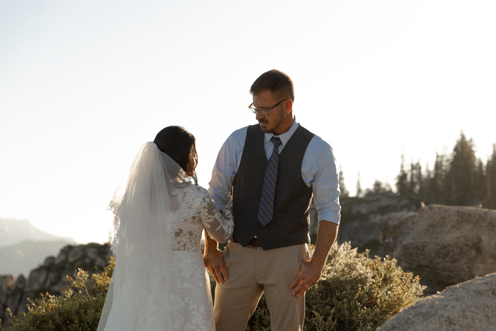Bride and groom standing outdoors in wedding attire, bride in lace dress and veil, groom in vest and tie, with a natural background of rocks and trees in soft lighting.
