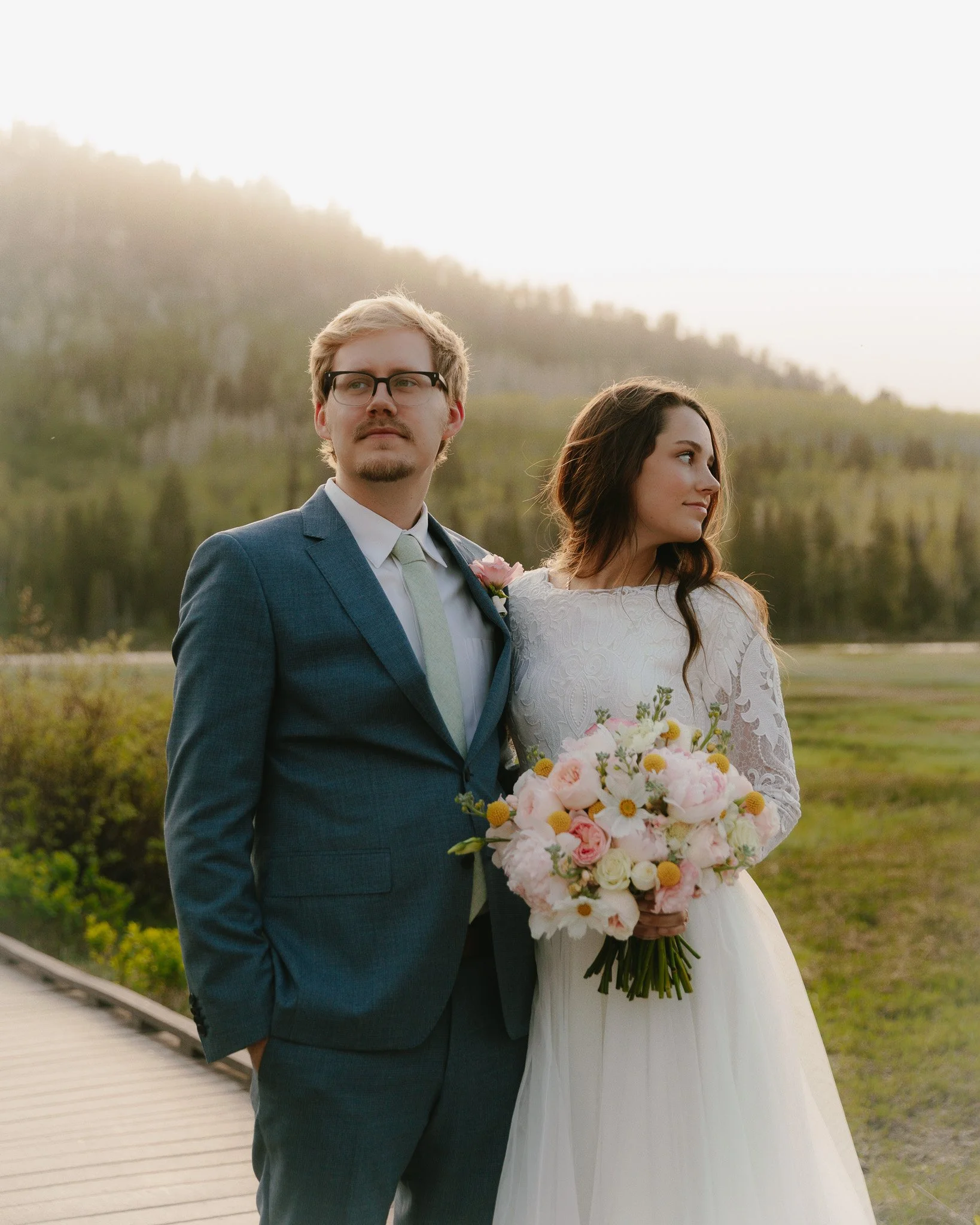 A groom in a blue suit and a bride in a white wedding dress standing outdoors on a wooden path, holding a bouquet of pink and white flowers, with a backdrop of green hills and trees.