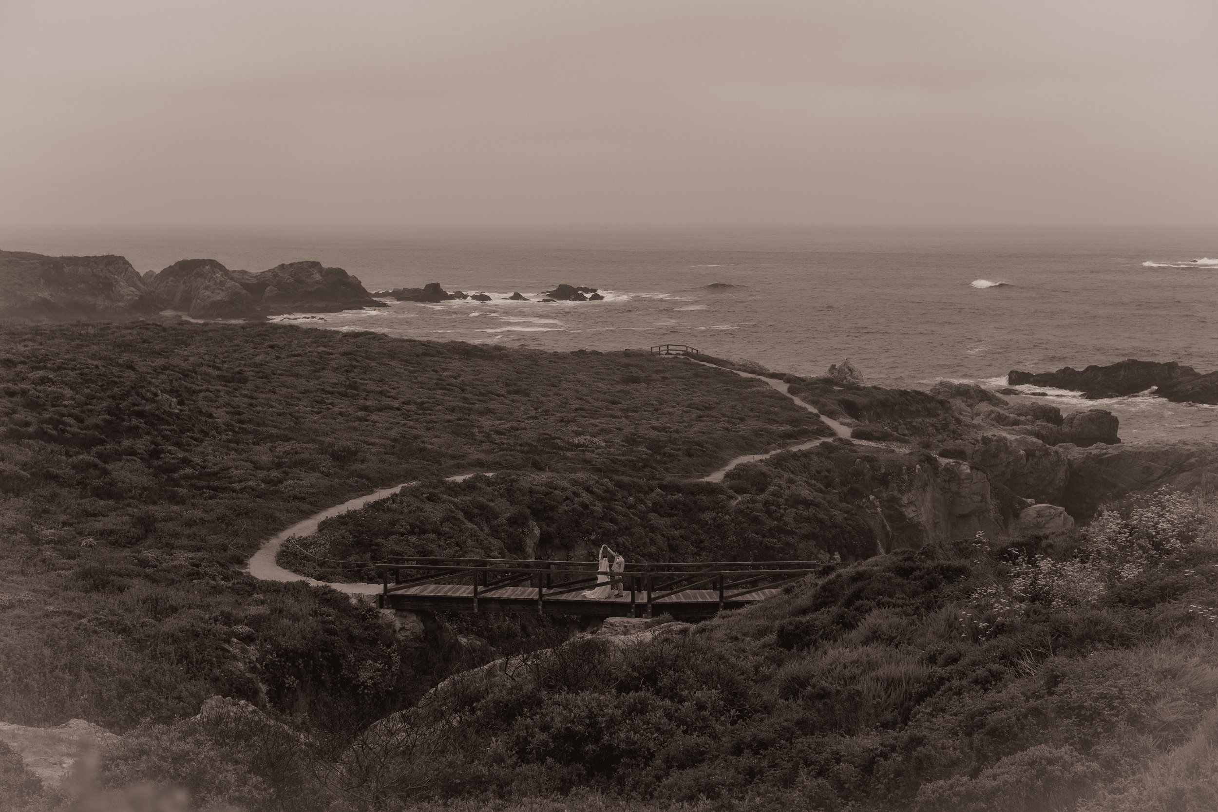 A couple standing on a wooden bridge overlooking a hiking trail along a rugged coastline with rocks and waves in the distance, in a monochrome sepia tone.